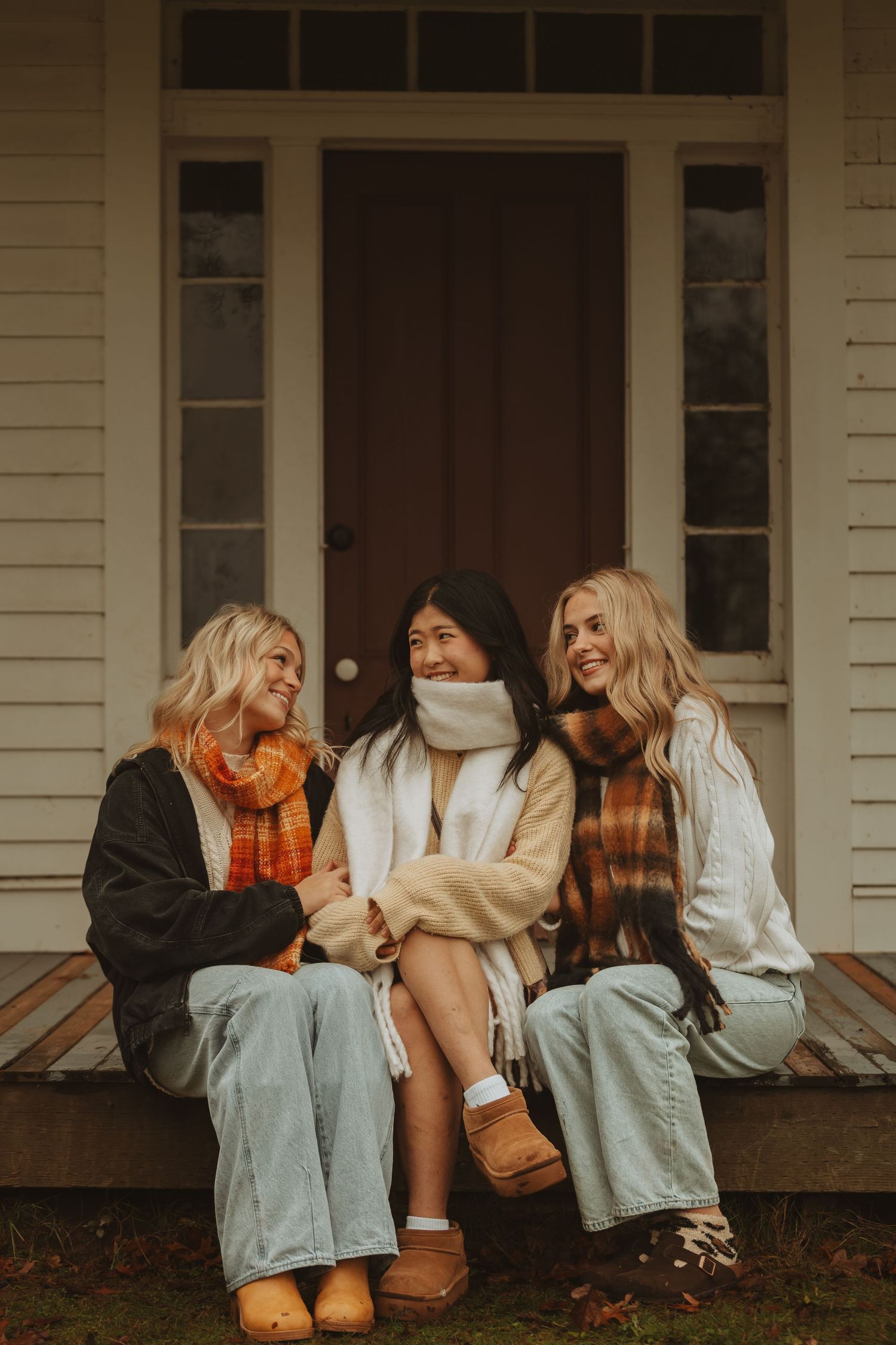 Four friends in casual attire sit and laugh together on a white porch during golden hour.