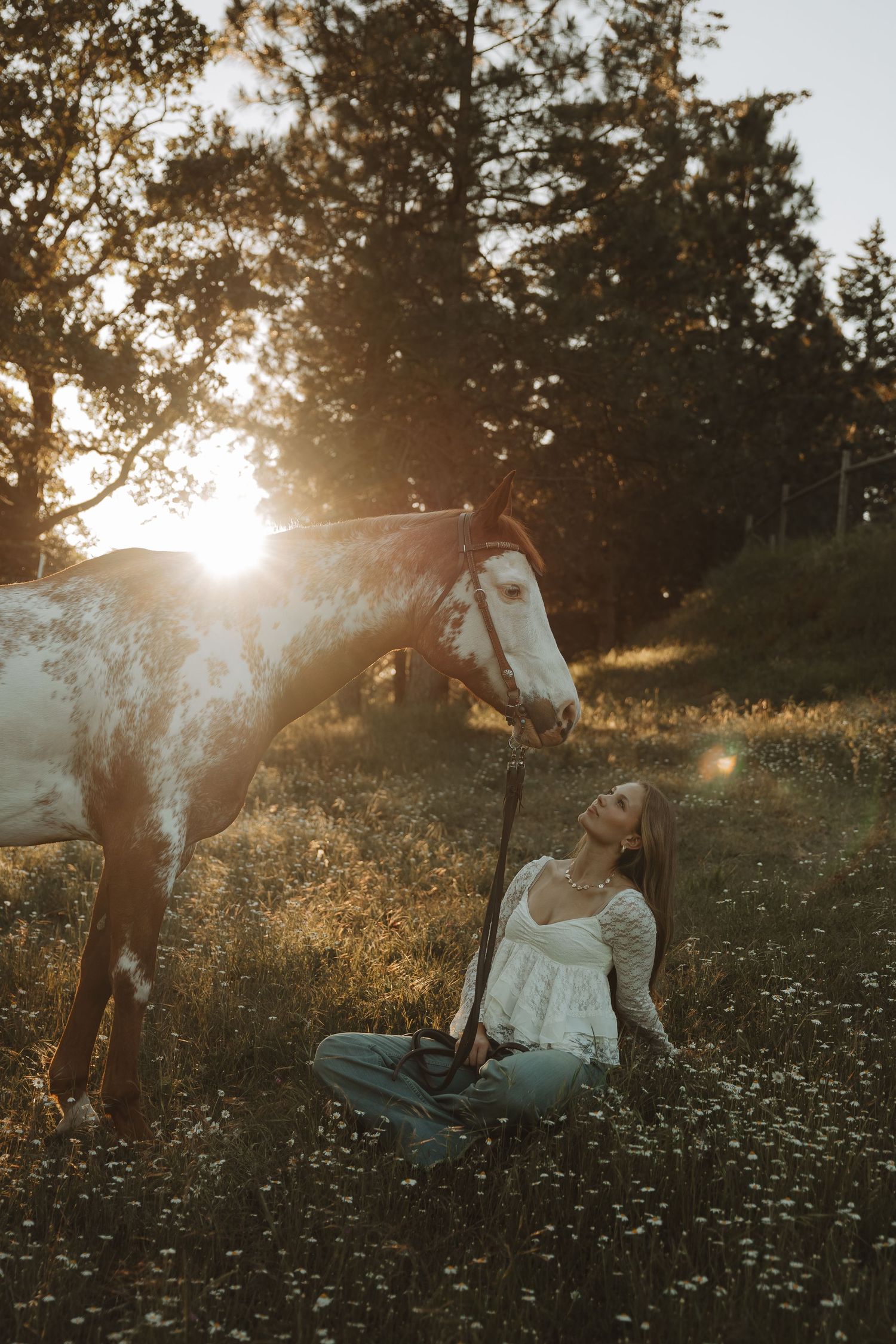 Woman in white top sits in wildflower field with appaloosa horse at golden hour sunset.