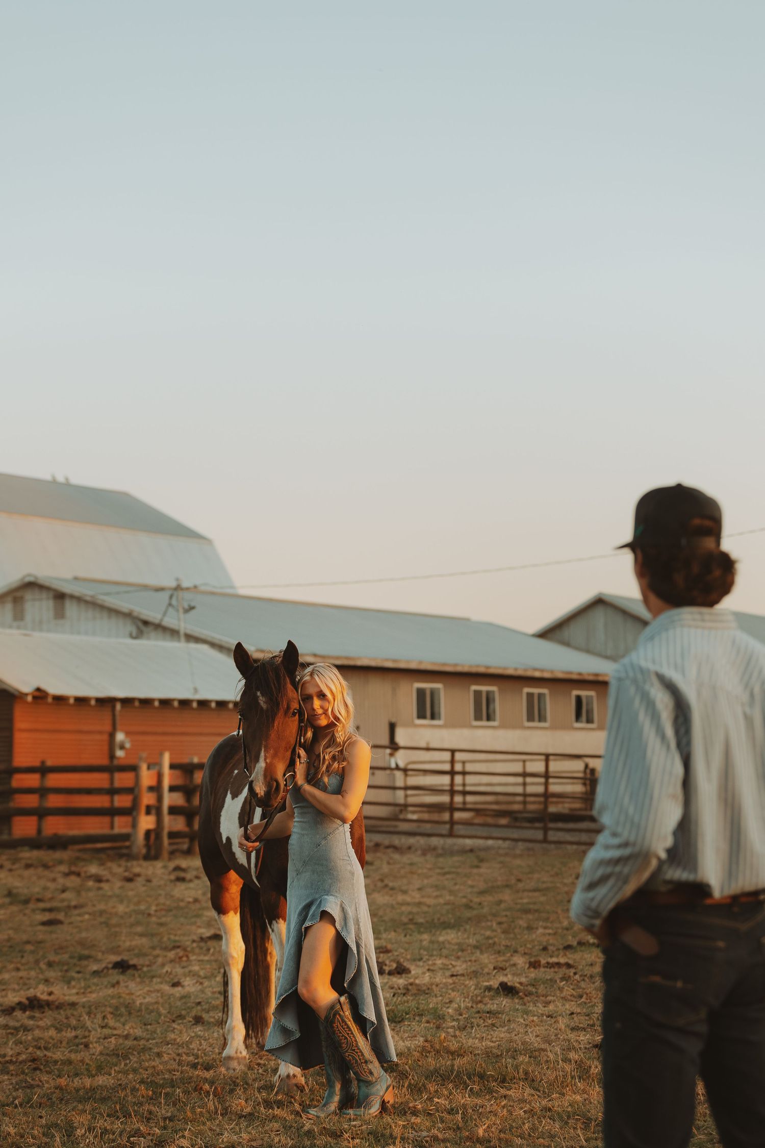 Woman in blue dress with brown horse at golden hour on a ranch, man in striped shirt watching nearby.