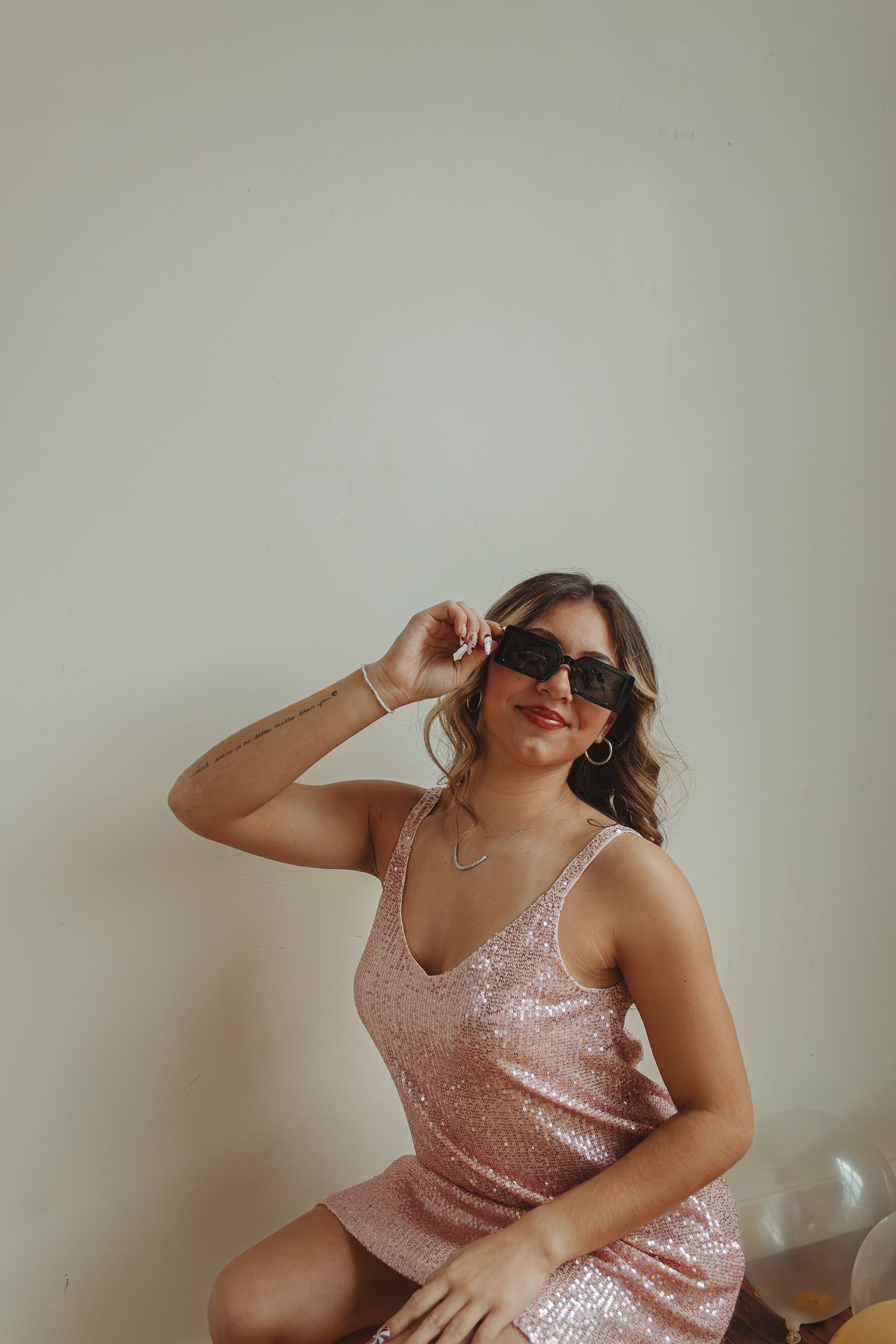 Person in sparkly pink dress poses playfully with sunglasses against neutral wall.