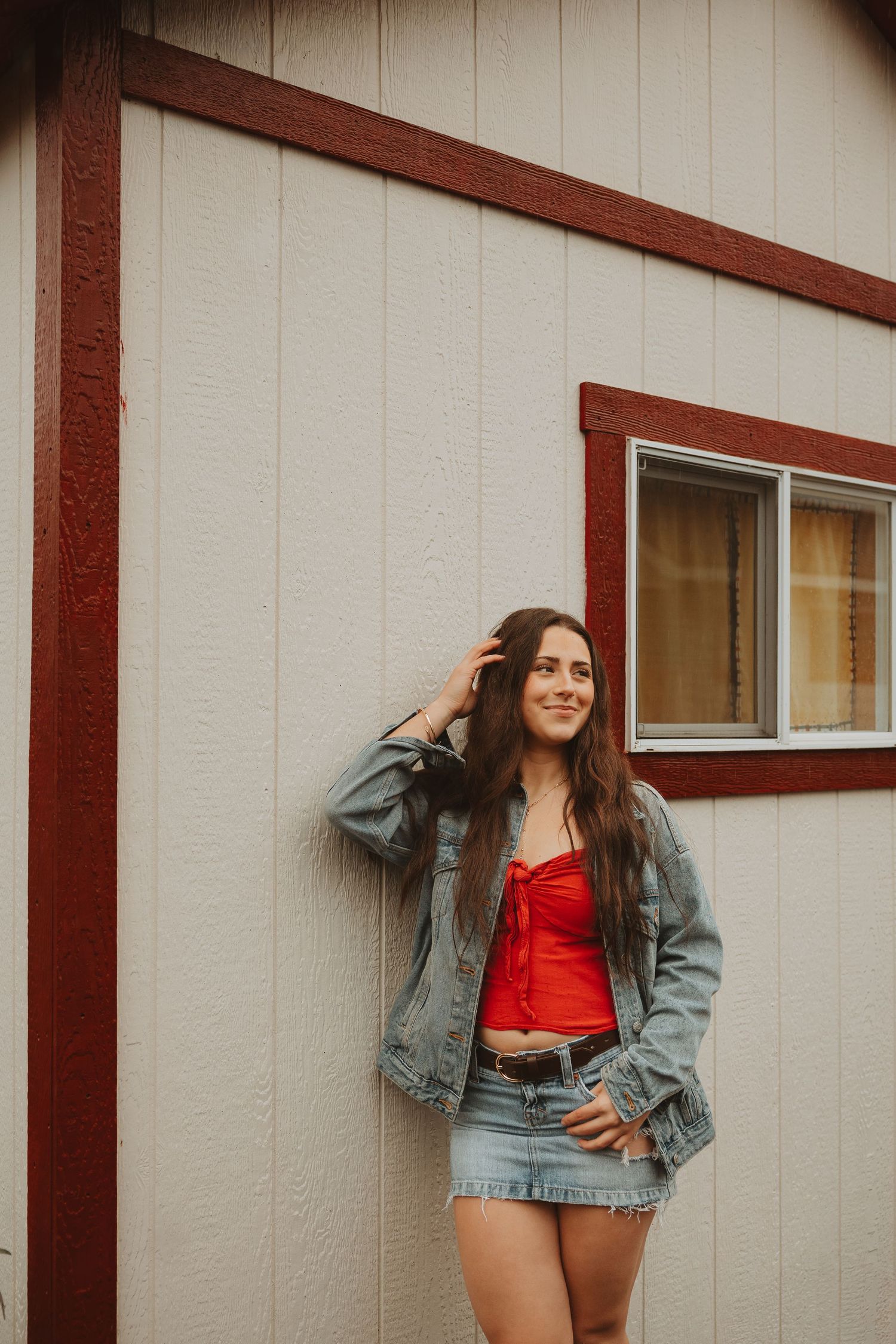 A casual pose against a white building with brown trim wearing denim jacket and red clothing.