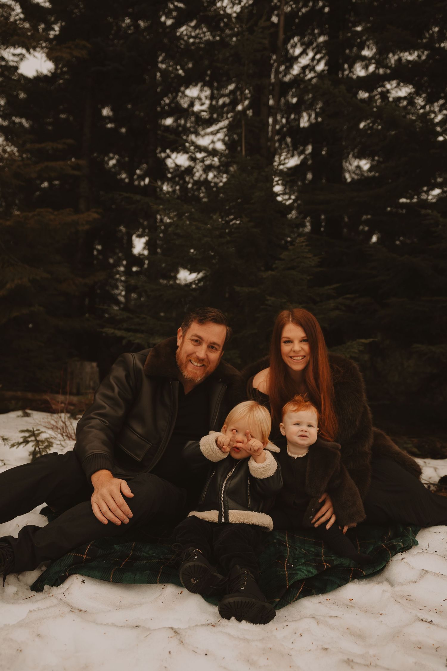 Family lying together on snowy ground surrounded by evergreen trees in a cozy winter setting.