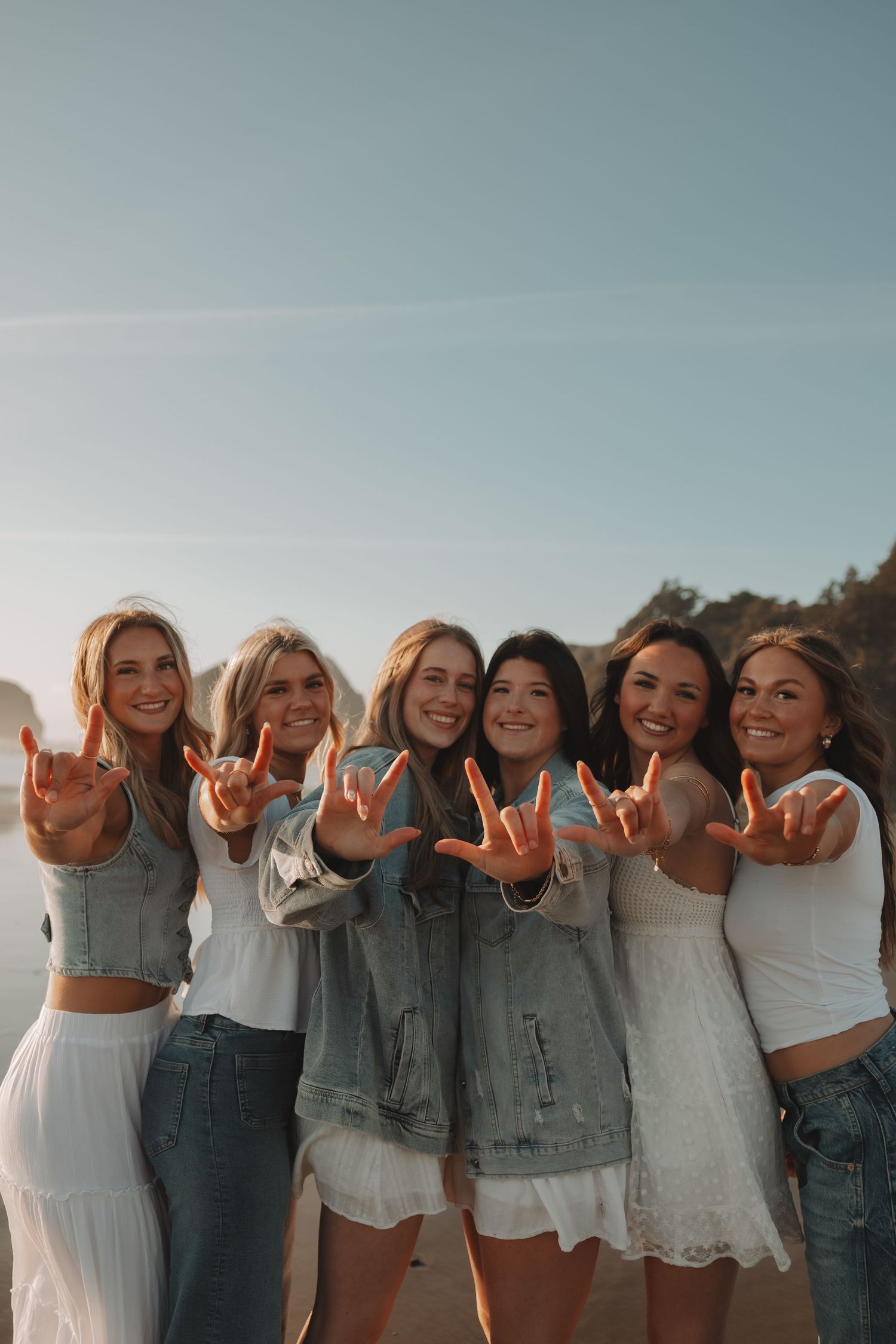 Six smiling young women at beach making shaka signs wearing white outfits and denim.