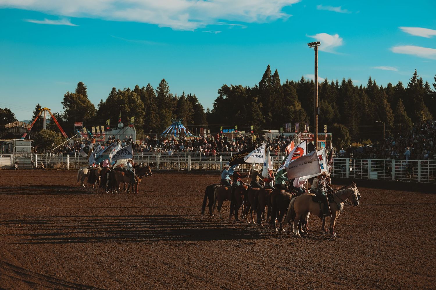 Panoramic view of a rodeo arena with blue sky, pine trees, and spectator stands in the background.