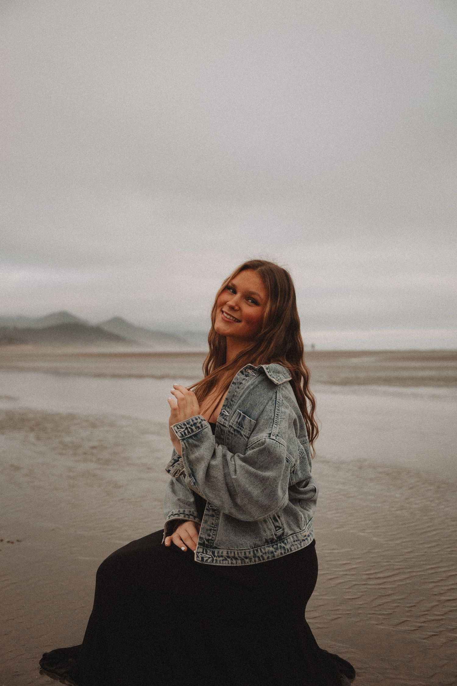 A joyful person in a denim jacket sits on a beach during an overcast day, laughing and enjoying the moment.