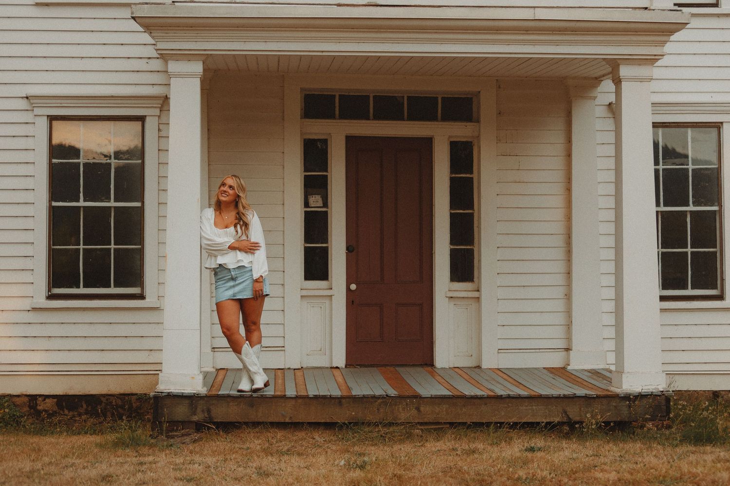 Young blonde woman in white top and denim skirt posing on porch of white farmhouse with brown door.