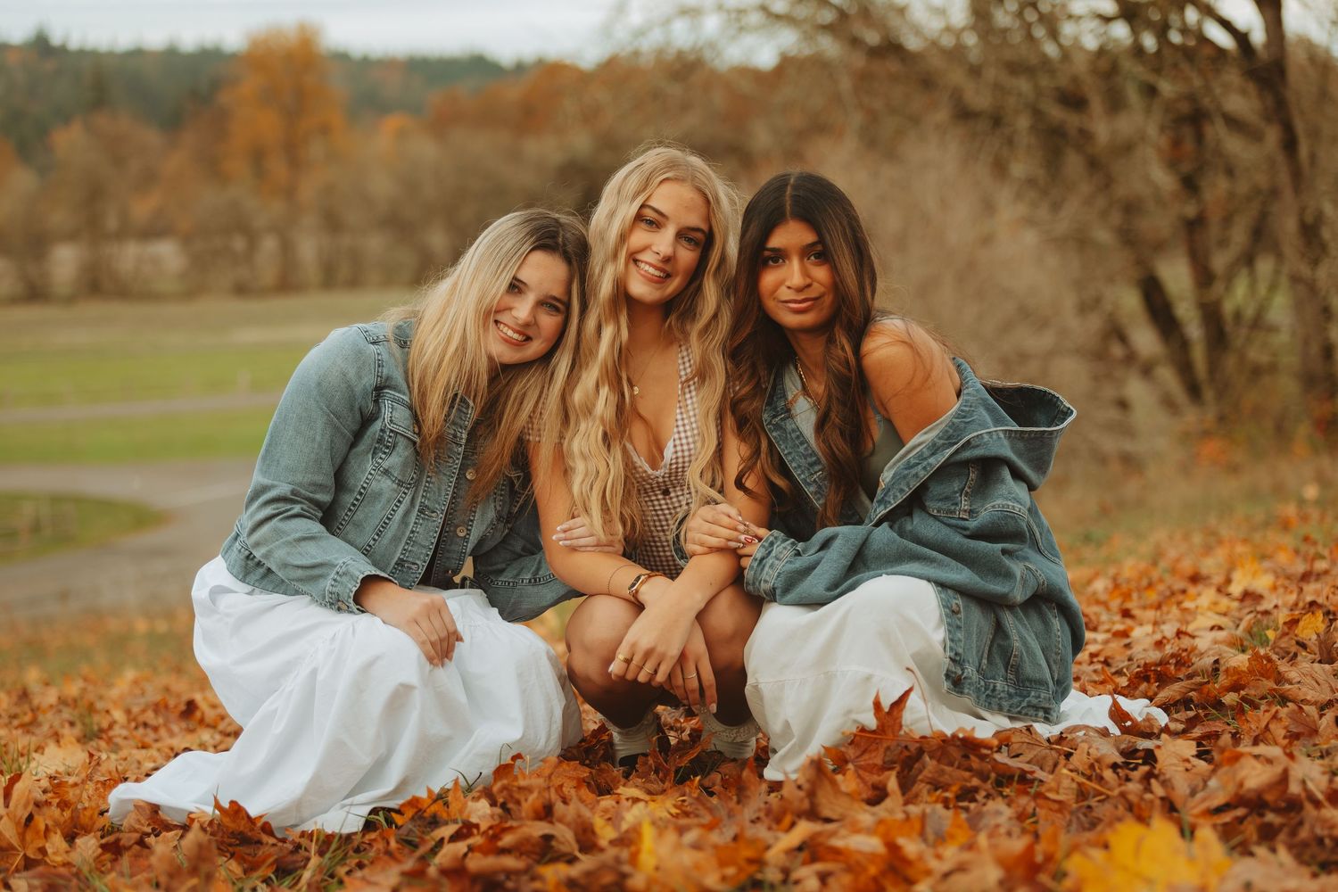 Friends lying together on autumn leaves wearing denim jackets and white dresses during a fall photoshoot.