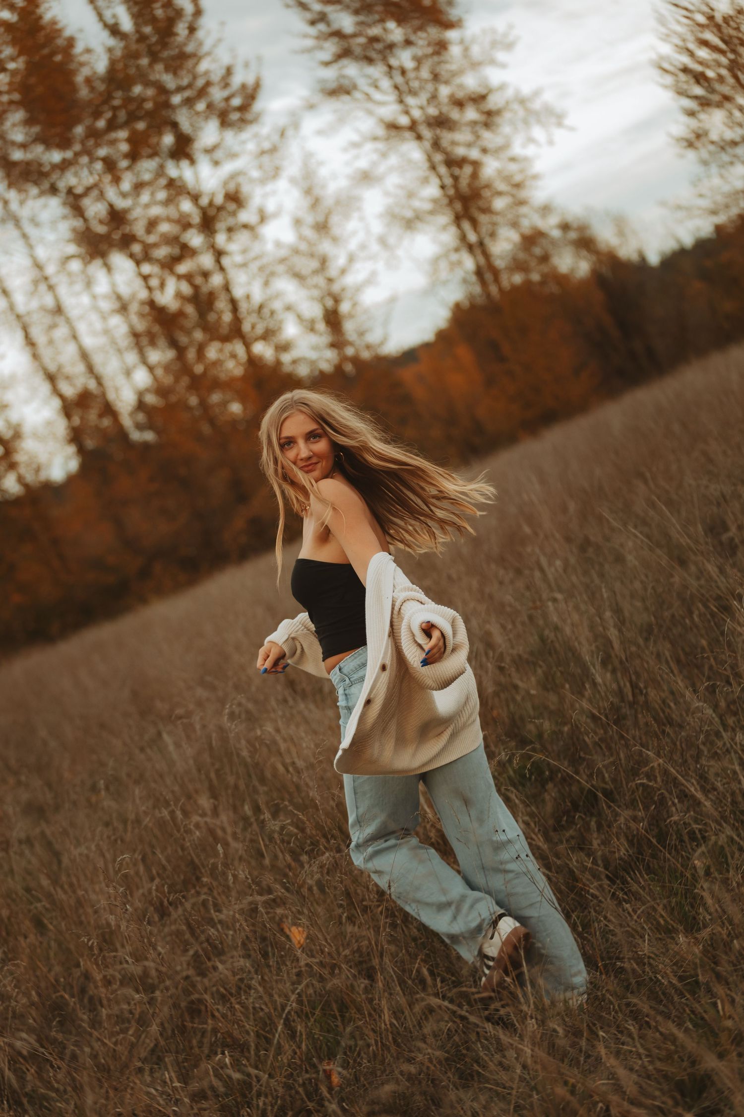 A woman in casual attire dances playfully in a rustic field with fall foliage in the background.