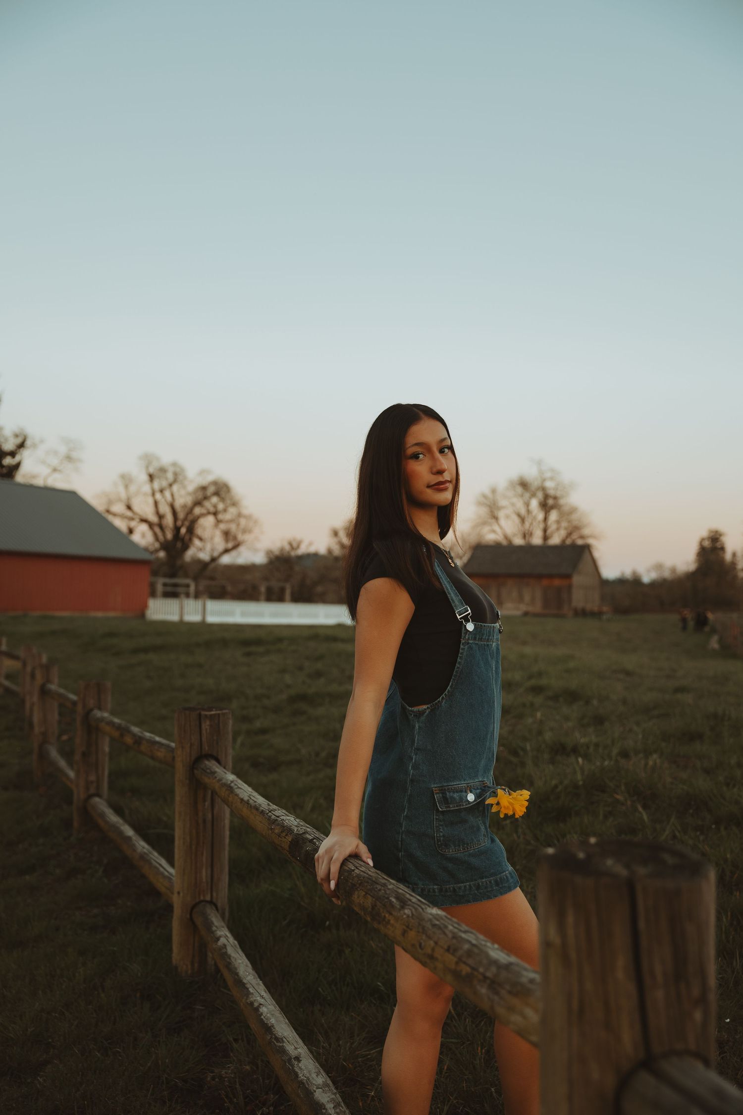Young woman in denim overalls poses by a wooden fence at a farm during sunset.