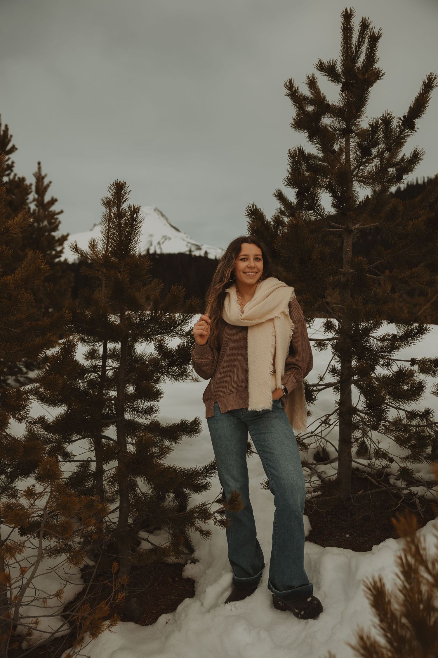 Woman in brown sweater and white scarf smiling in snowy mountain forest with pine trees and peak behind her.