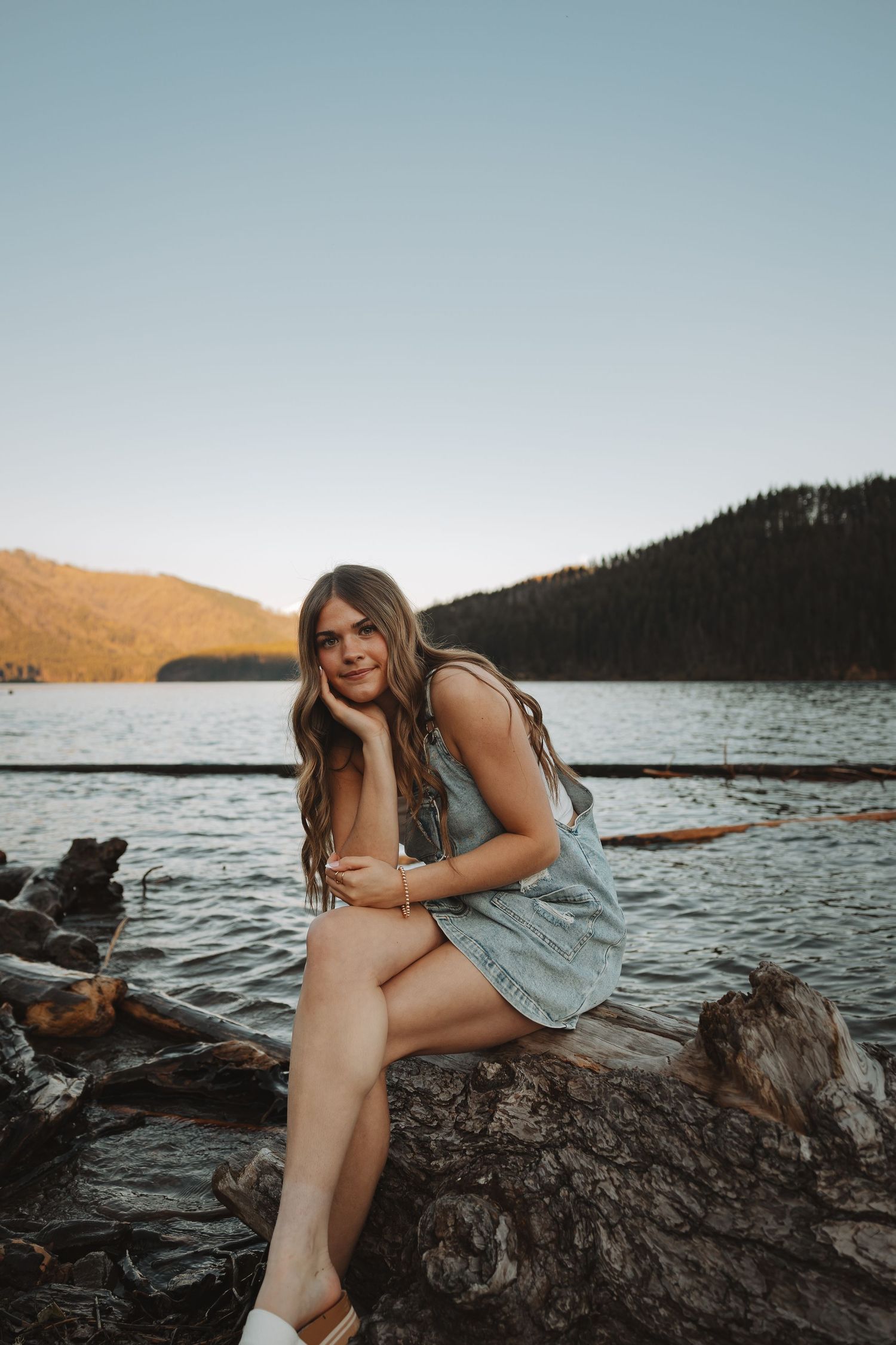 Young woman in denim dress sitting on driftwood log by mountain lake at golden hour sunset.