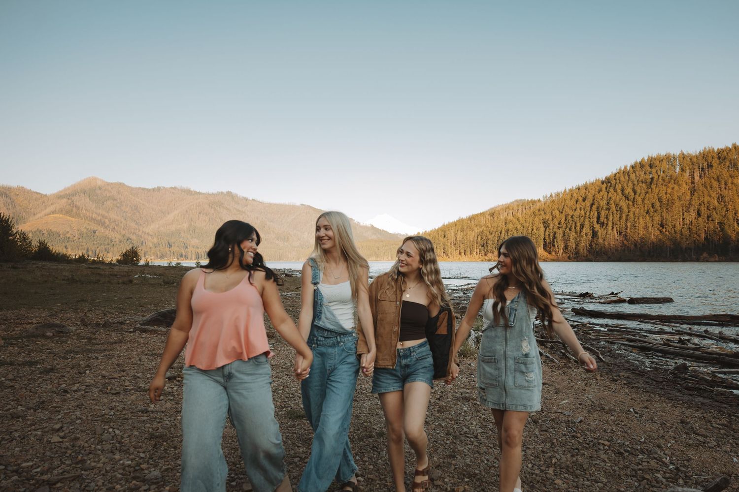 Four young women holding hands walking along a rocky lakeside beach with mountains and trees at sunset.