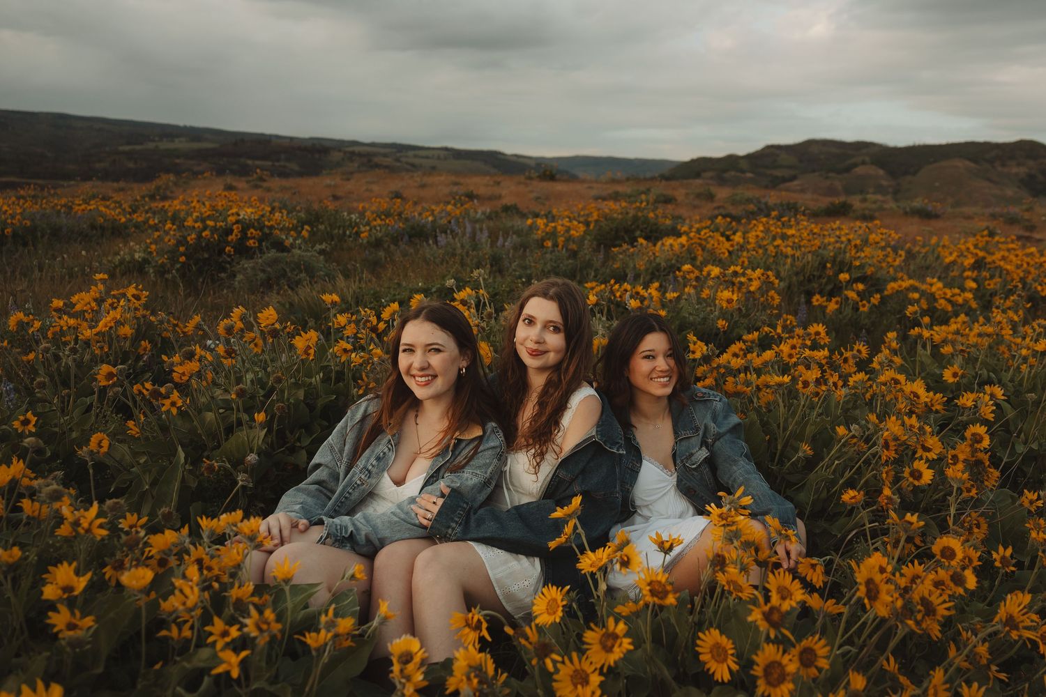 Three women sit smiling in a vast field of yellow wildflowers under a cloudy sky.