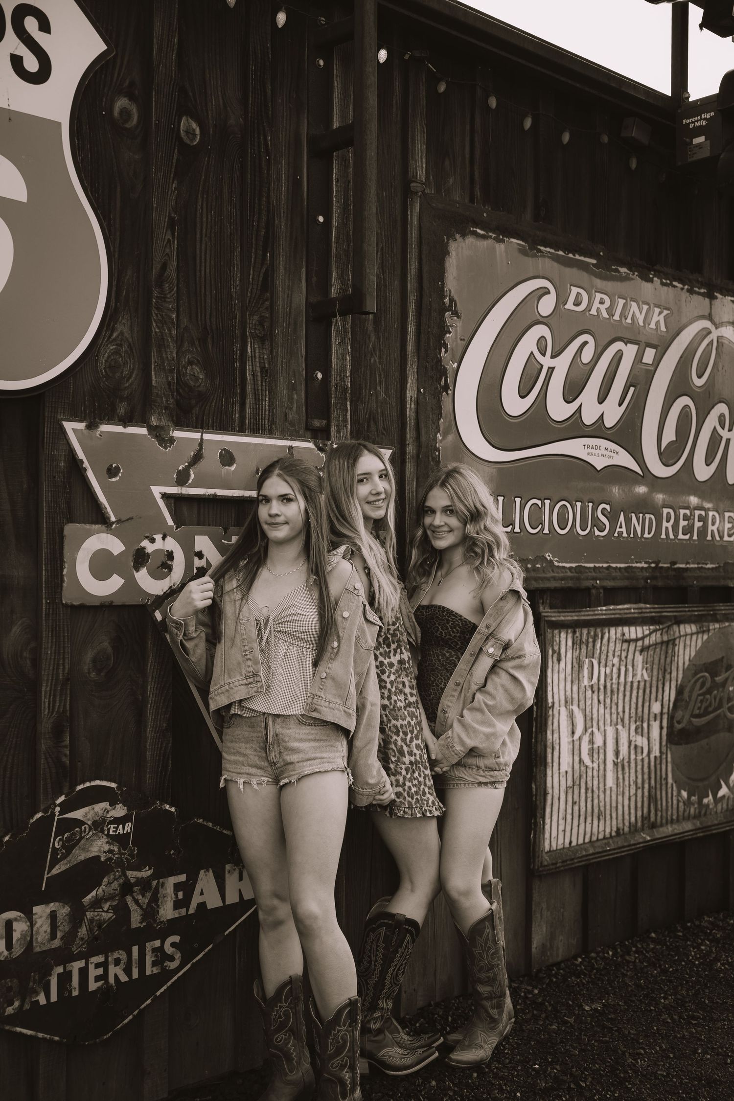 Three young women in cowboy boots pose by vintage Coca-Cola and Goodyear signs on a rustic wooden wall.