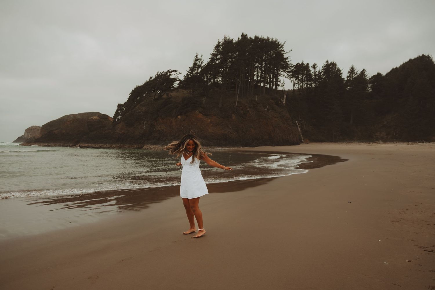 Woman in white dress standing on a sandy beach with rocky cliffs and trees under an overcast sky.