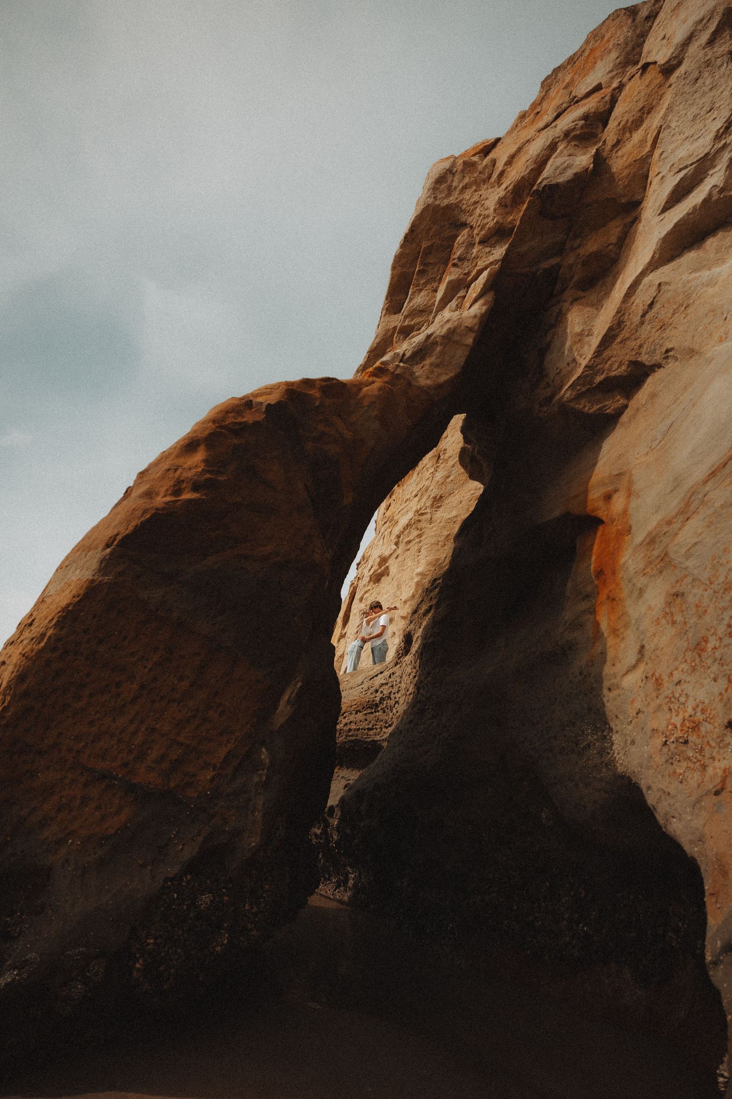 Couple embraces in narrow sandstone rock arch opening under moody sky.