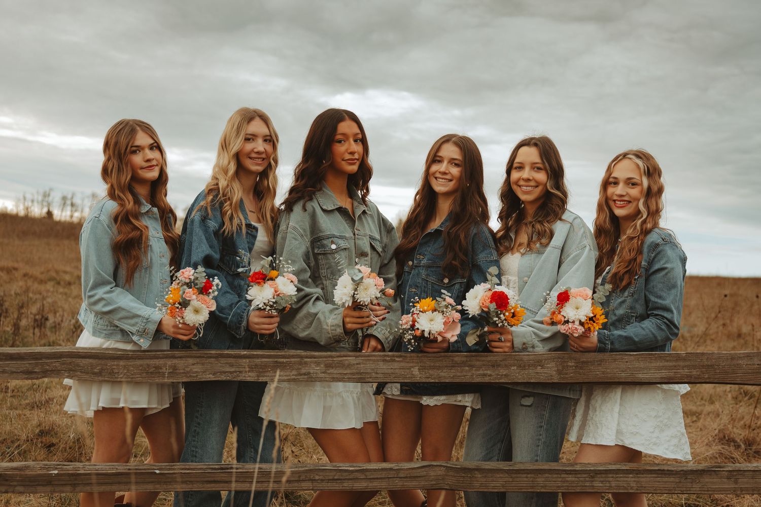 Bridesmaids in matching denim jackets pose together holding colorful floral bouquets against a cloudy sky.