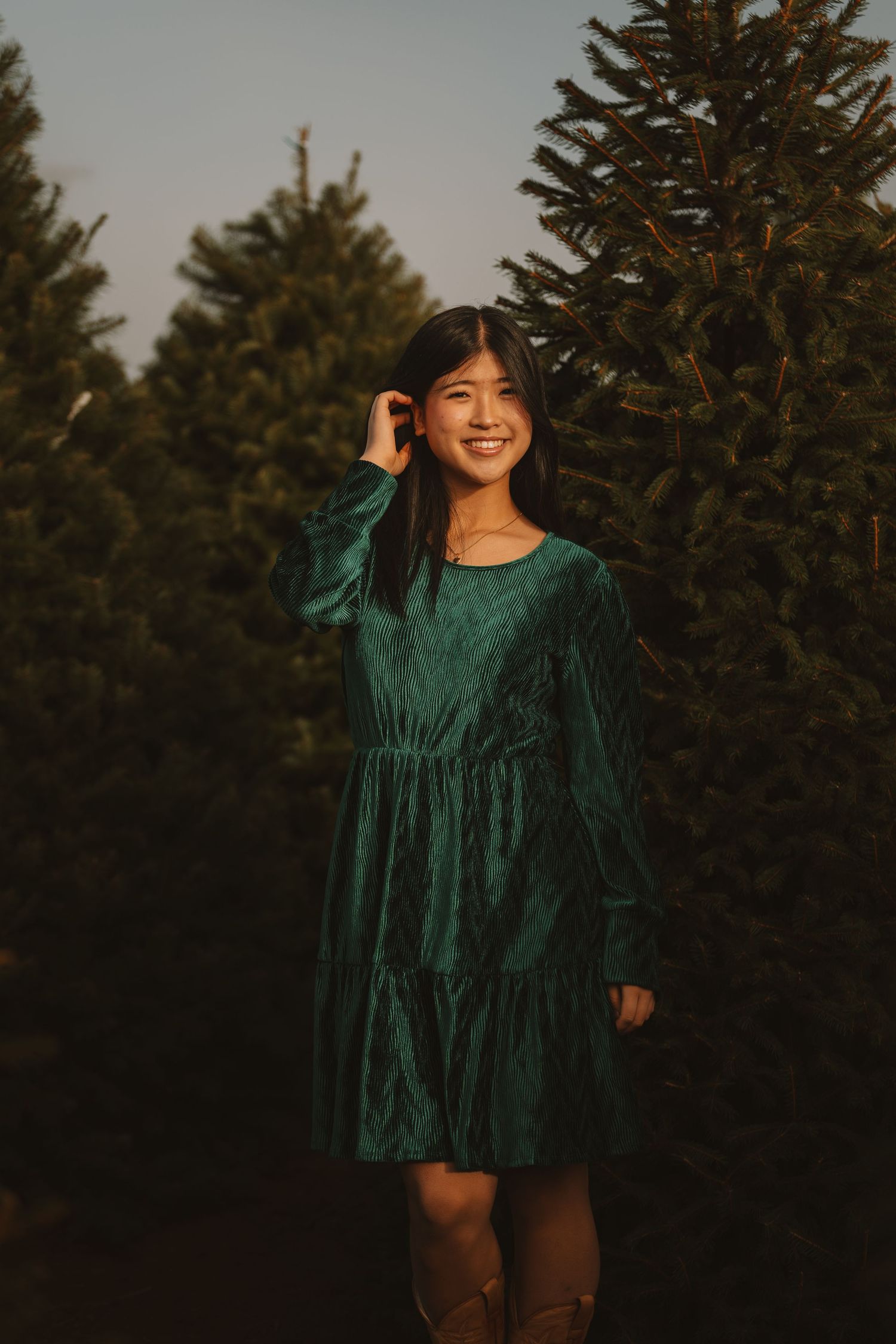 Person in emerald velvet dress poses among evergreen trees at sunset.