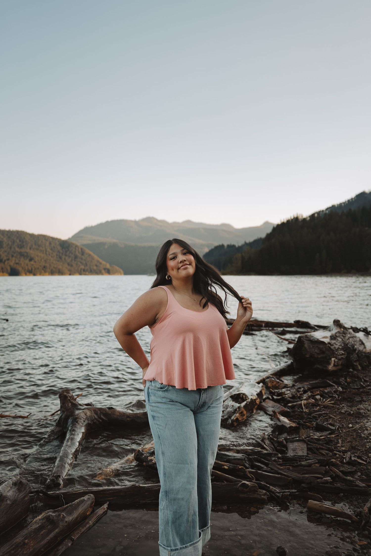 Smiling woman in pink tank top and jeans poses on driftwood logs by a scenic mountain lake.
