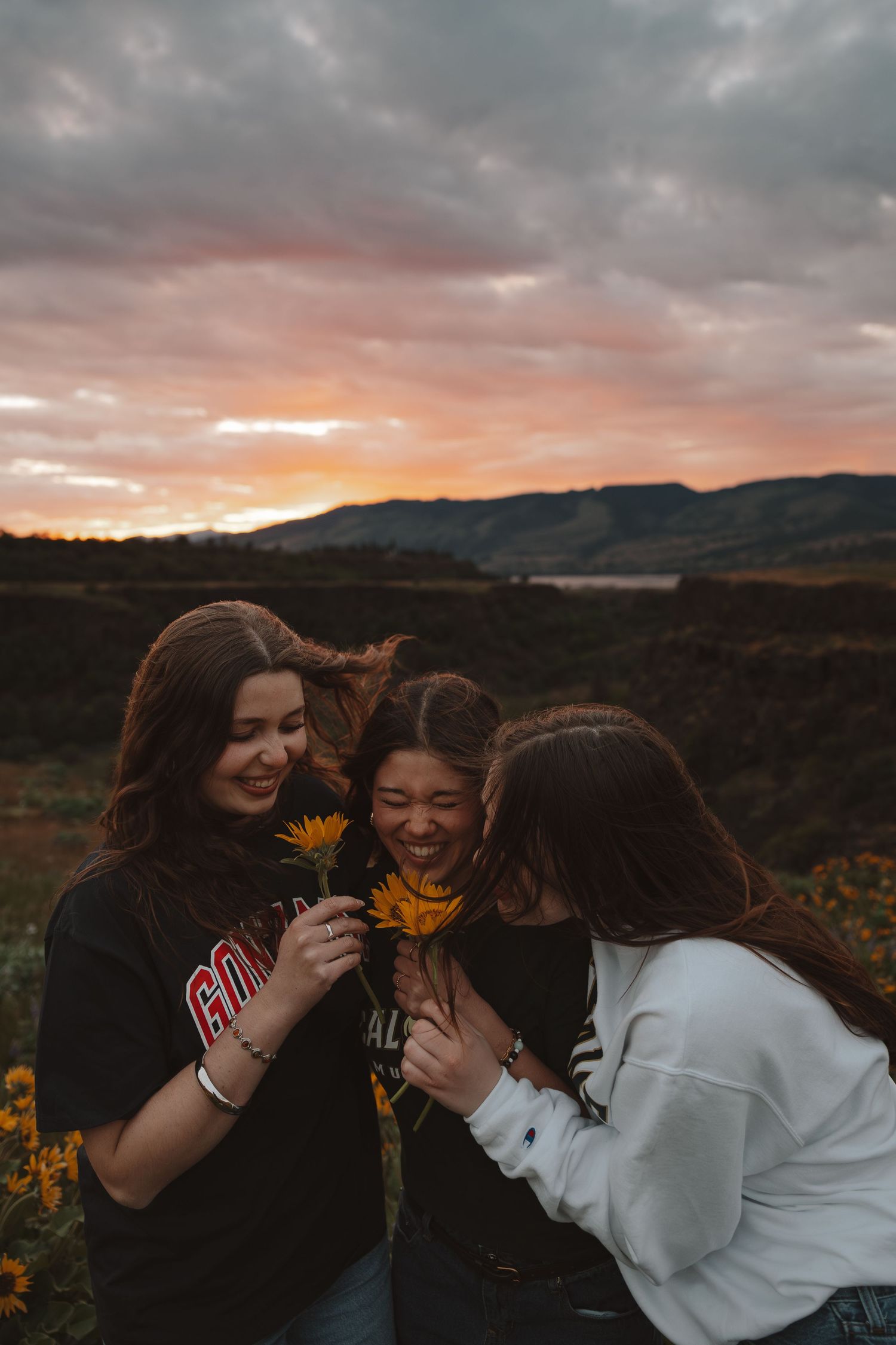 Three young women smiling and sharing a sunflower in a field during a vibrant sunset.