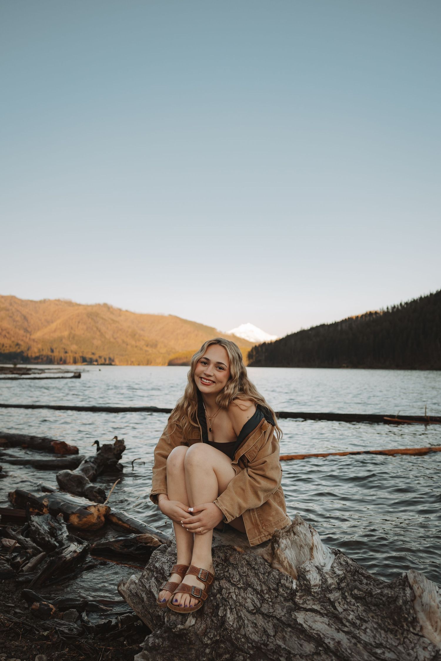 Smiling blonde woman sitting on rocks by a serene lake with mountains and golden sunset in the background.