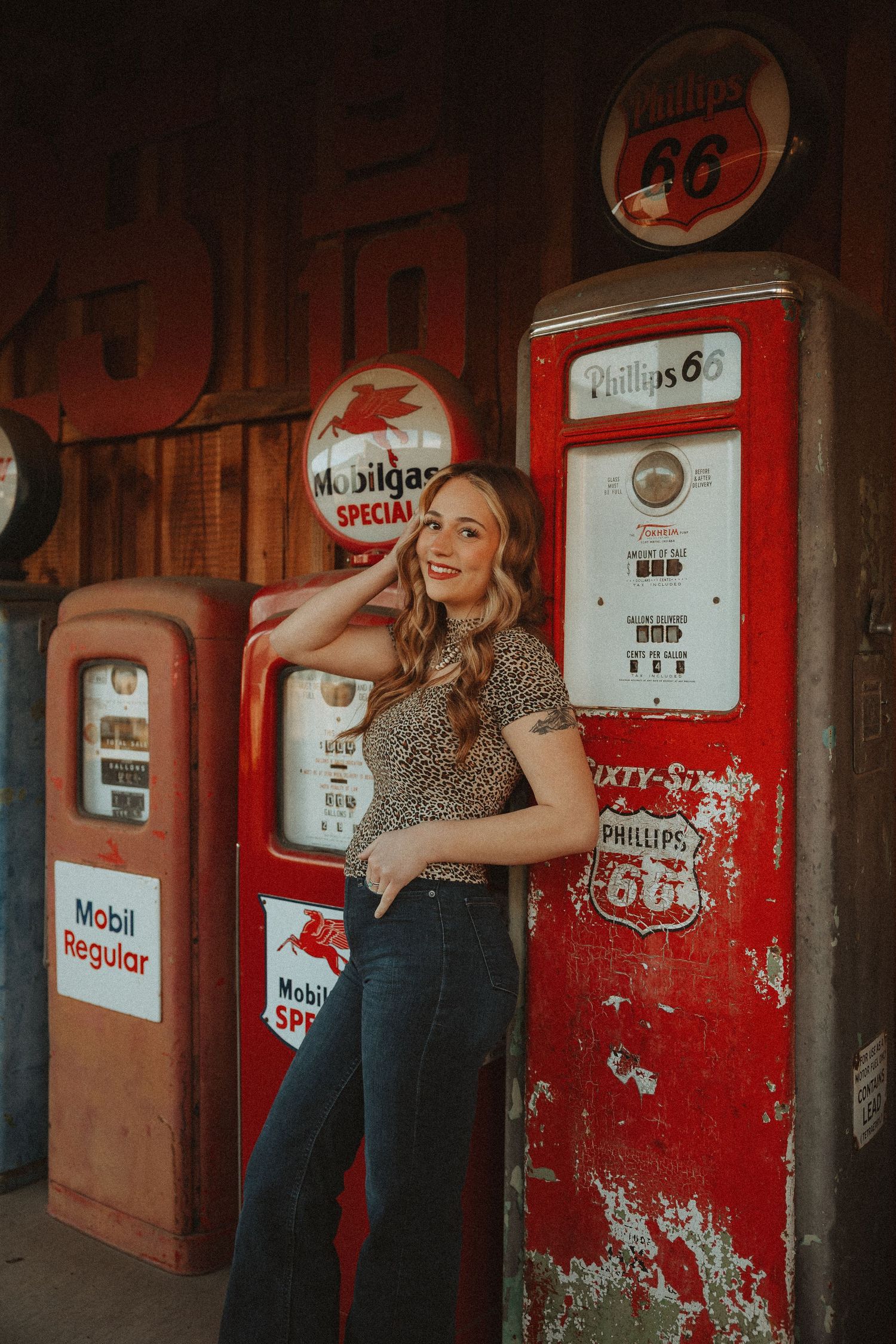 Young woman smiling in leopard print top and jeans beside vintage Phillips 66 and Mobilgas gas pumps.