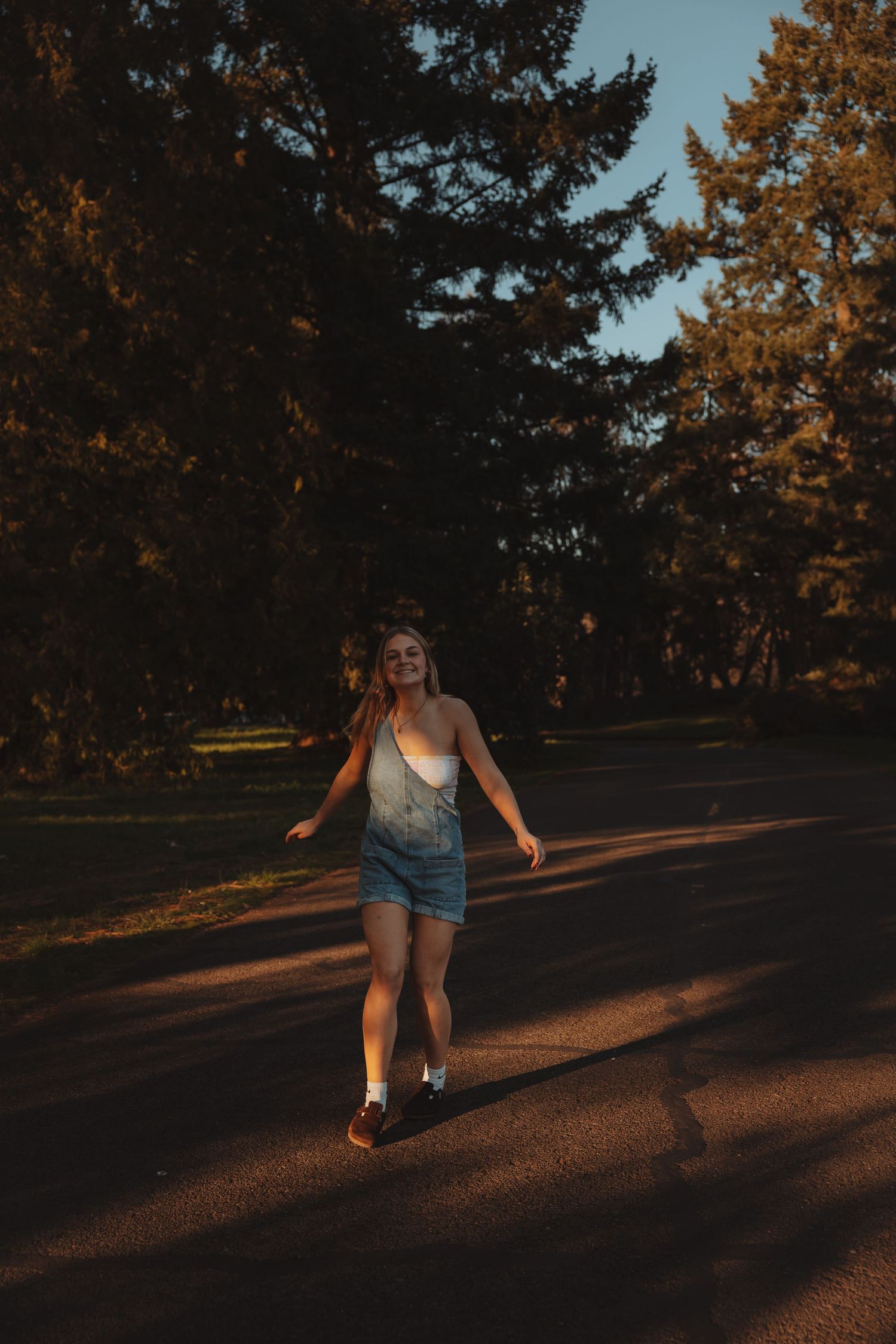 Young woman in denim overalls walking on a sunlit park road surrounded by tall trees at golden hour.