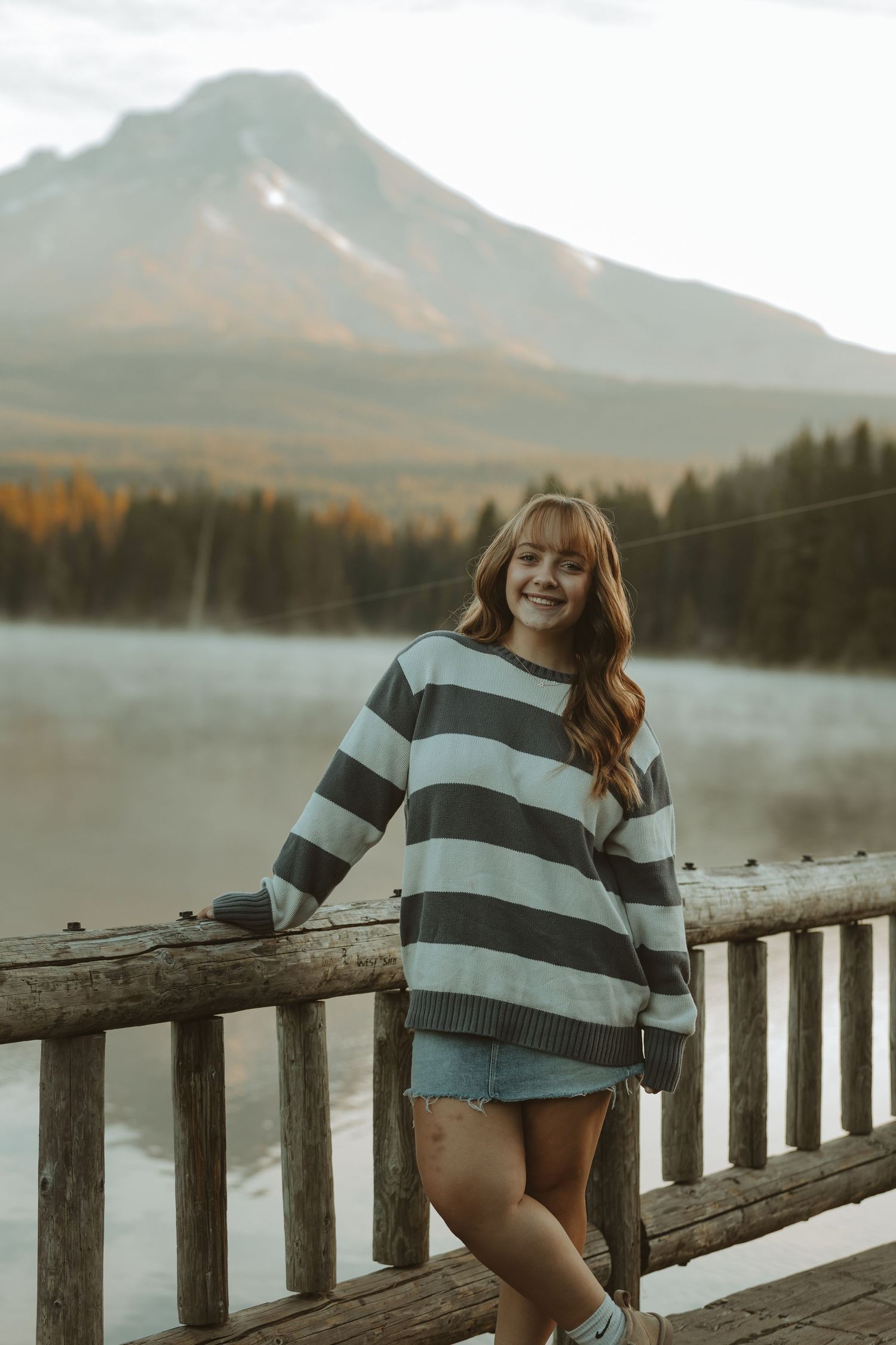 Person in striped sweater poses by wooden railing at misty mountain lake during autumn sunset.