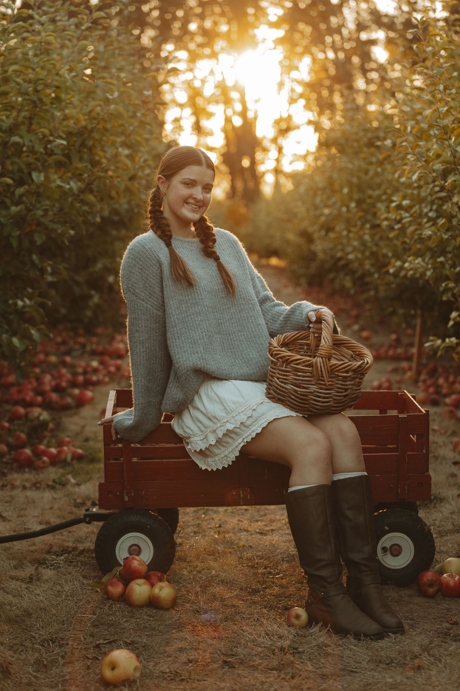 Person sits on rustic red wagon with basket and apples at sunset in an autumn orchard.