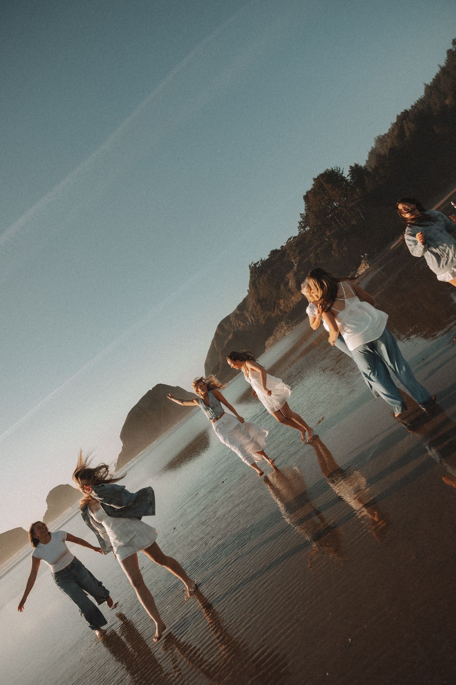 Group of friends dancing joyfully on a sunlit beach with rocky cliffs in the background at golden hour.