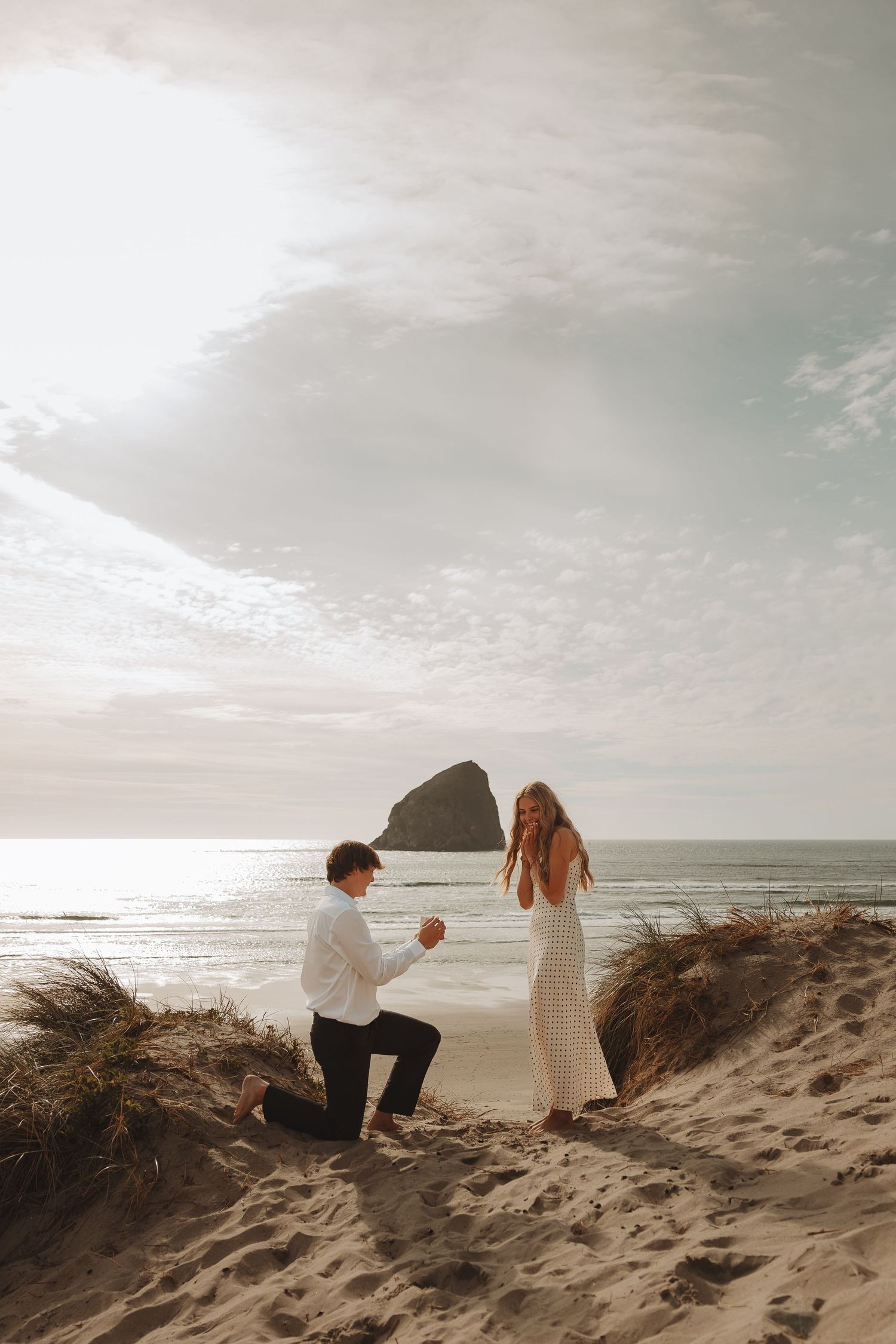 Couple shares romantic beach proposal moment on sand dunes with ocean and rock formation behind.