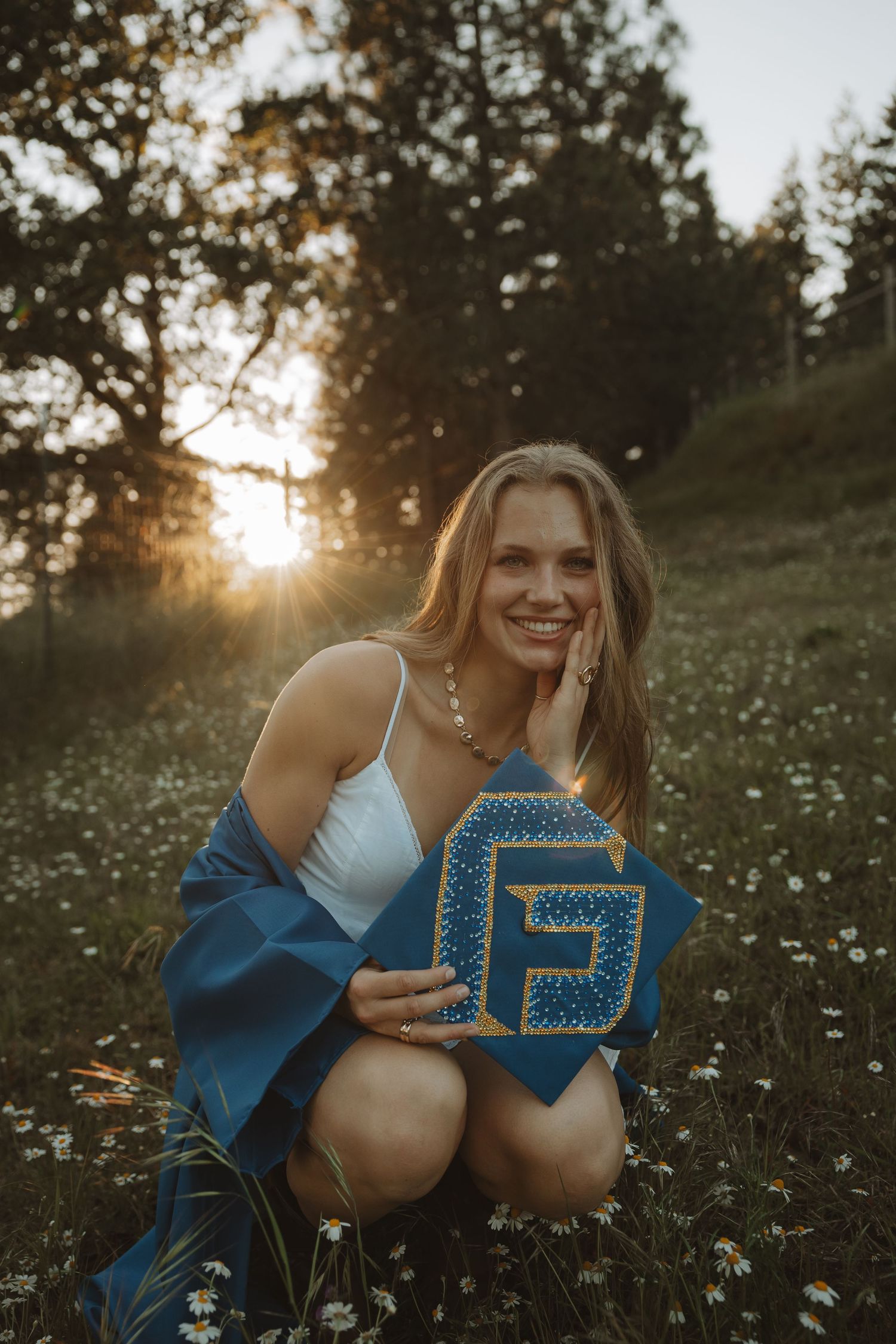 Smiling blonde graduate crouches in wildflower meadow holding decorated blue graduation cap at sunset.