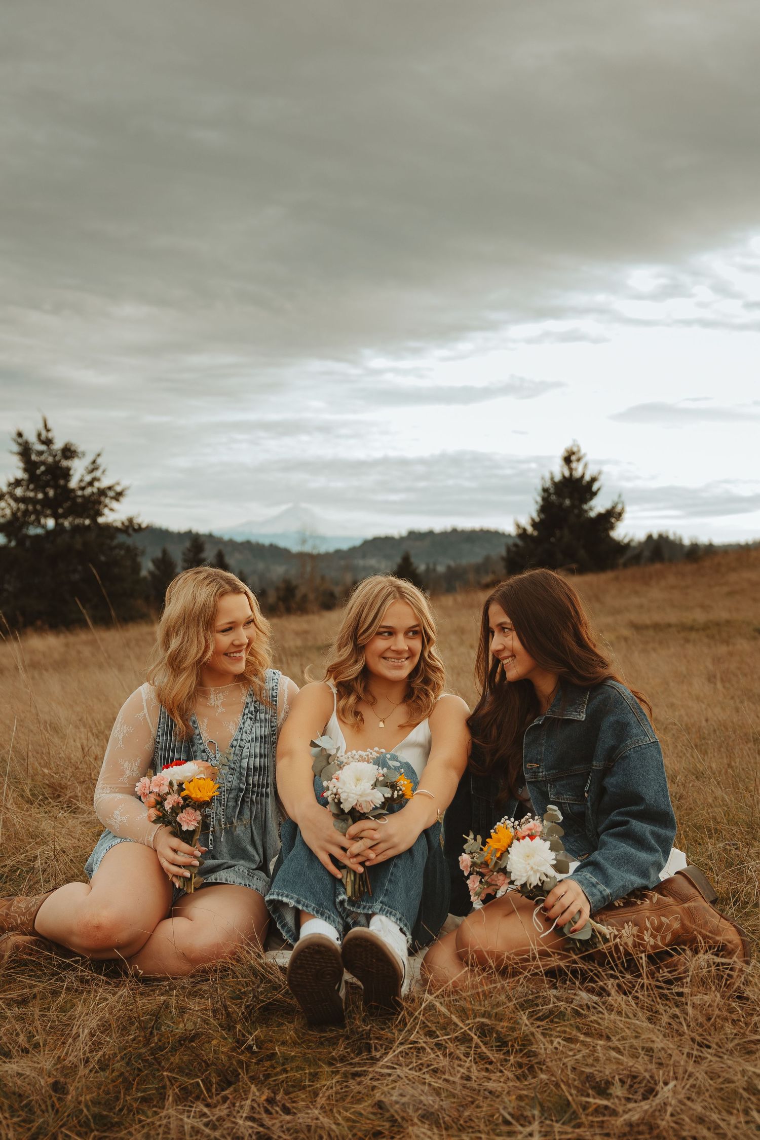 Three friends in denim jackets sit casually in grass holding colorful bouquets while laughing together.