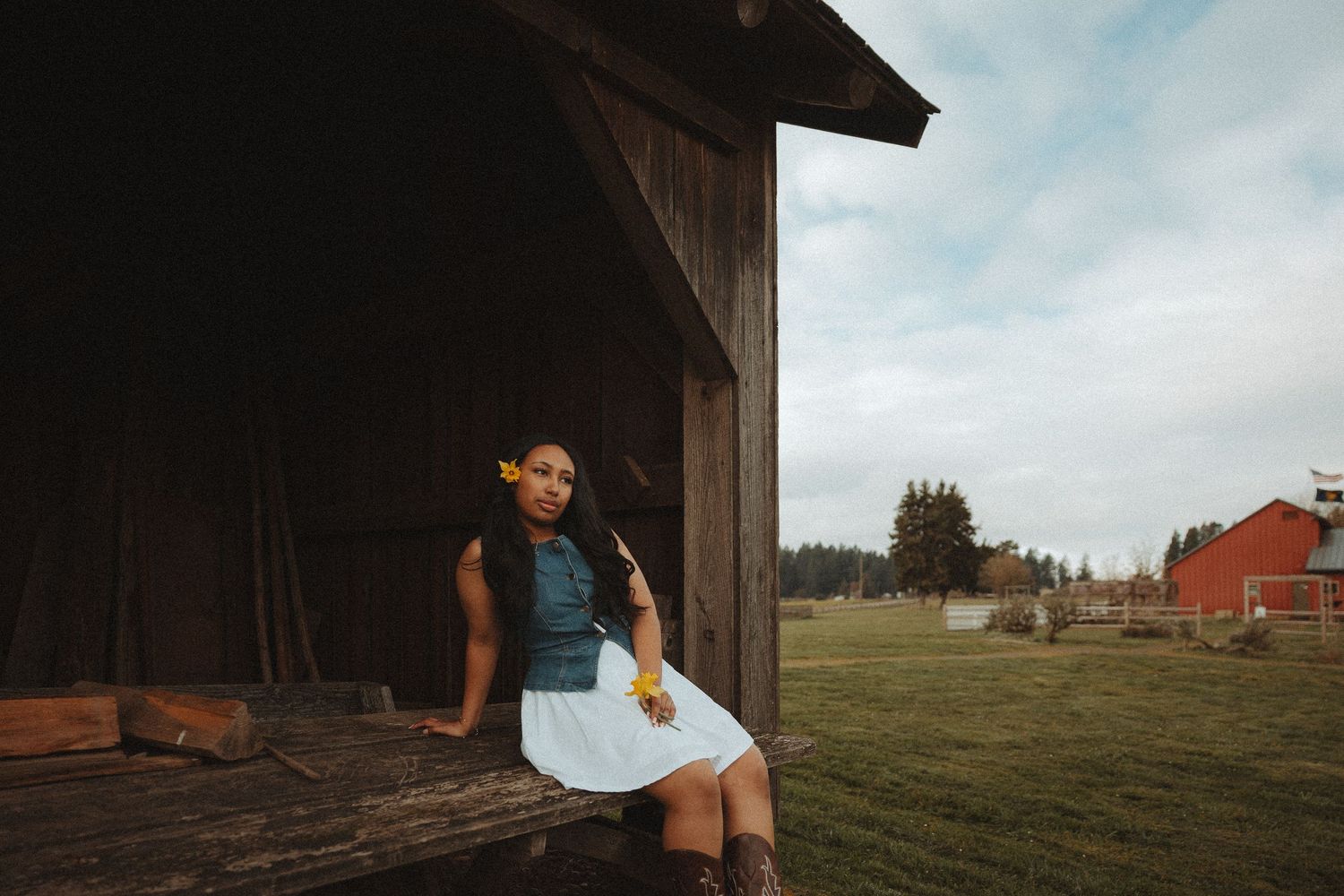 A rustic barn setting with someone sitting on the edge wearing a white dress on a sunny summer day with hay bales in the field.