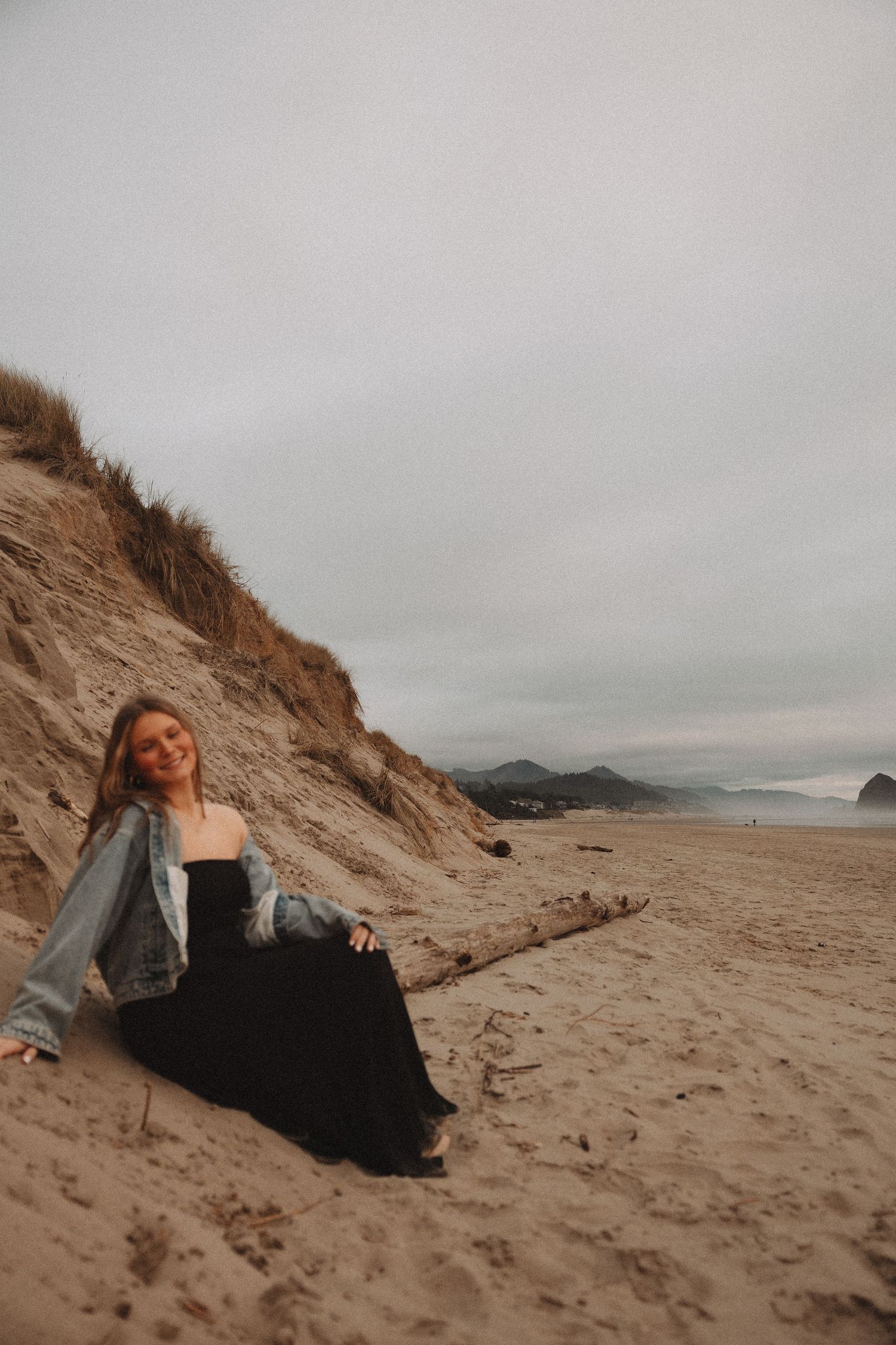 Person sitting on sandy beach at sunset with dramatic coastal cliffs and ocean view in background.