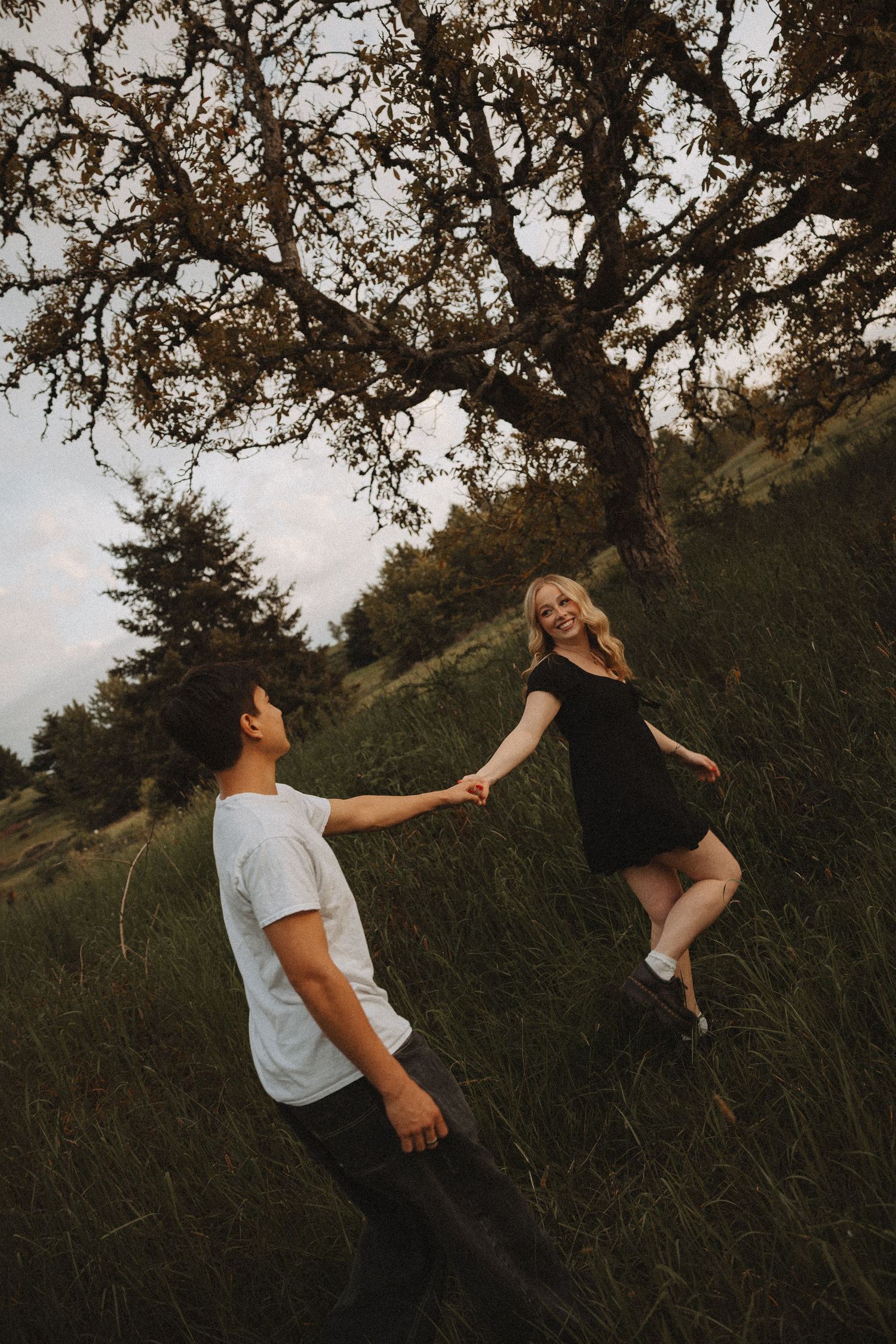 Couple holding hands in a grassy field with a large tree, woman in black dress smiling at man in white shirt.