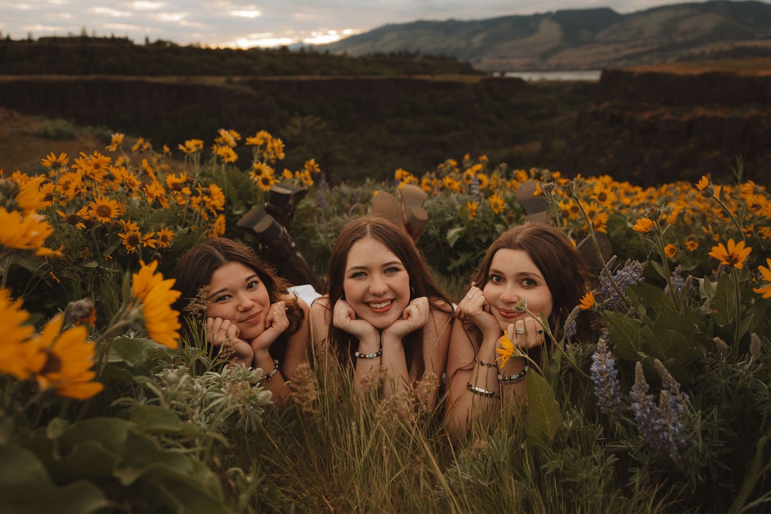 Three smiling young women lying in a vibrant yellow wildflower field with mountains in the background.