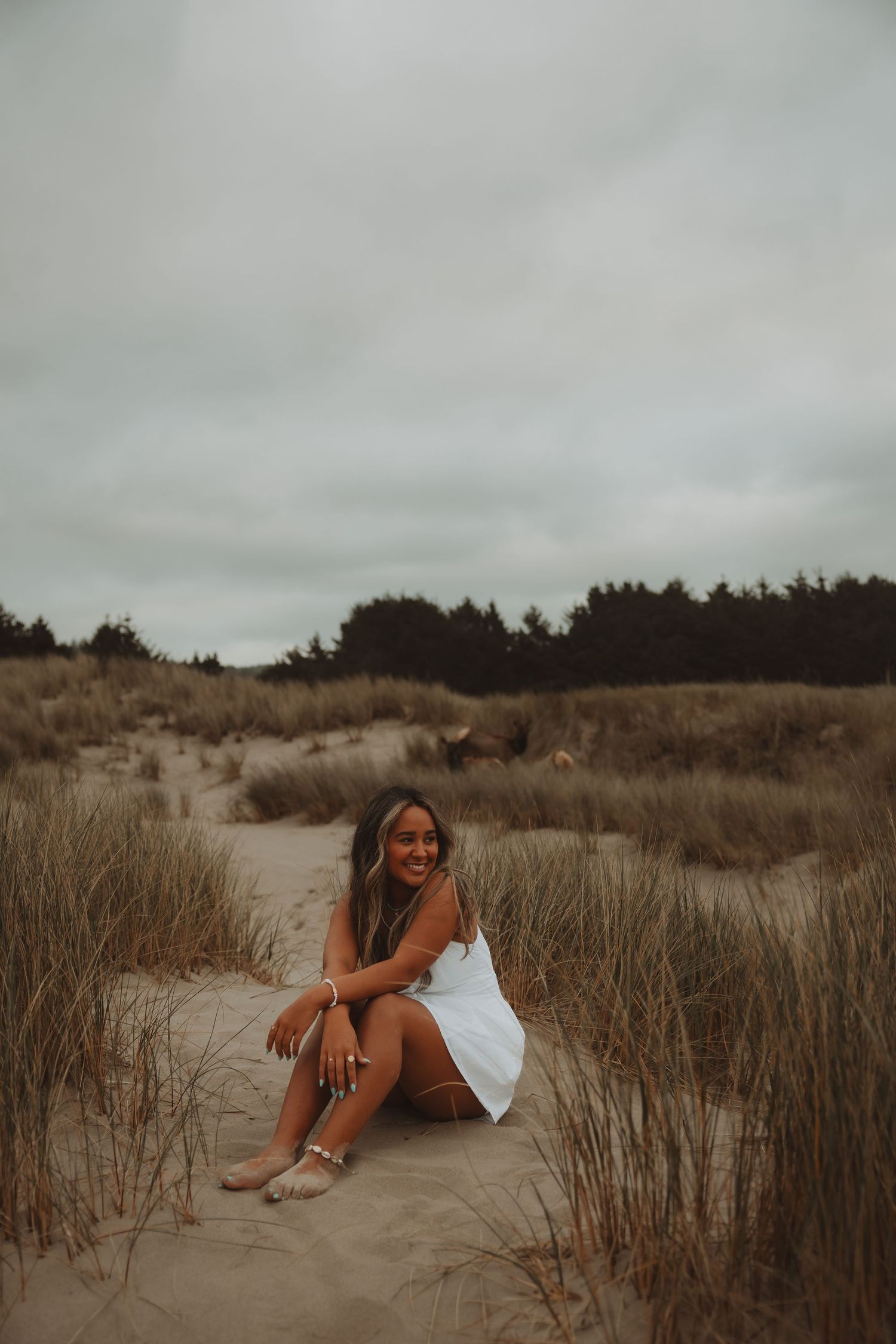 Young woman in white dress sitting on sandy beach dunes with dry grass and trees on overcast day.