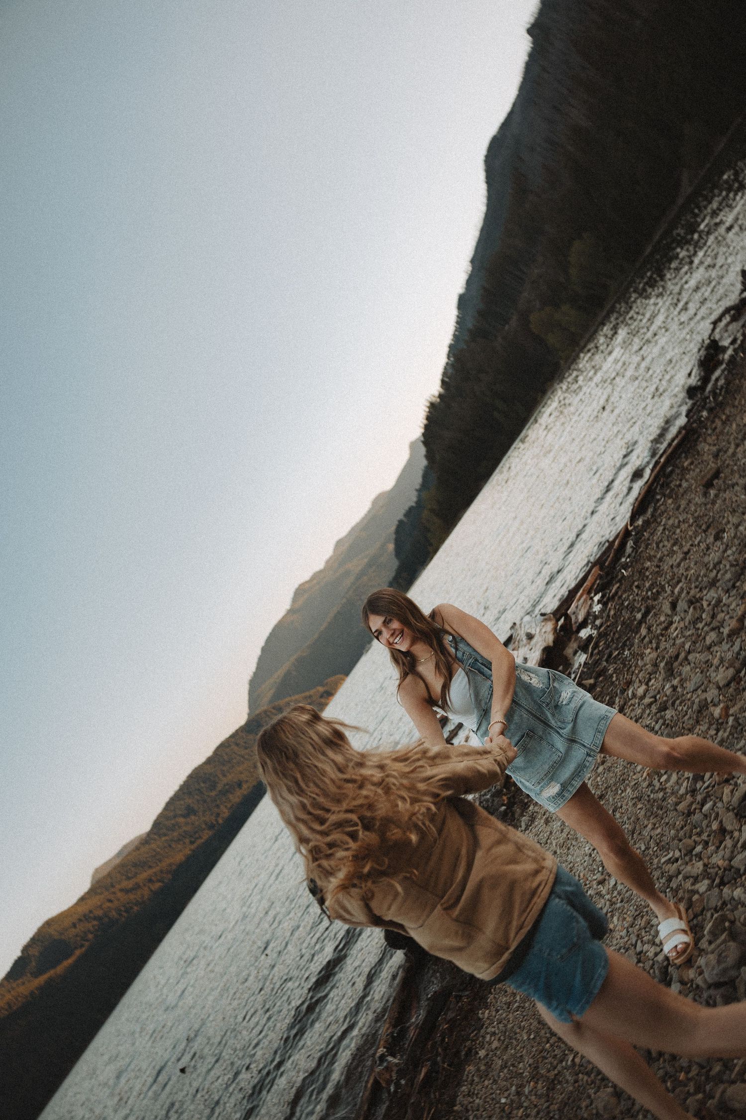 Two young women laughing and holding hands on a rocky shoreline with mountain backdrop.
