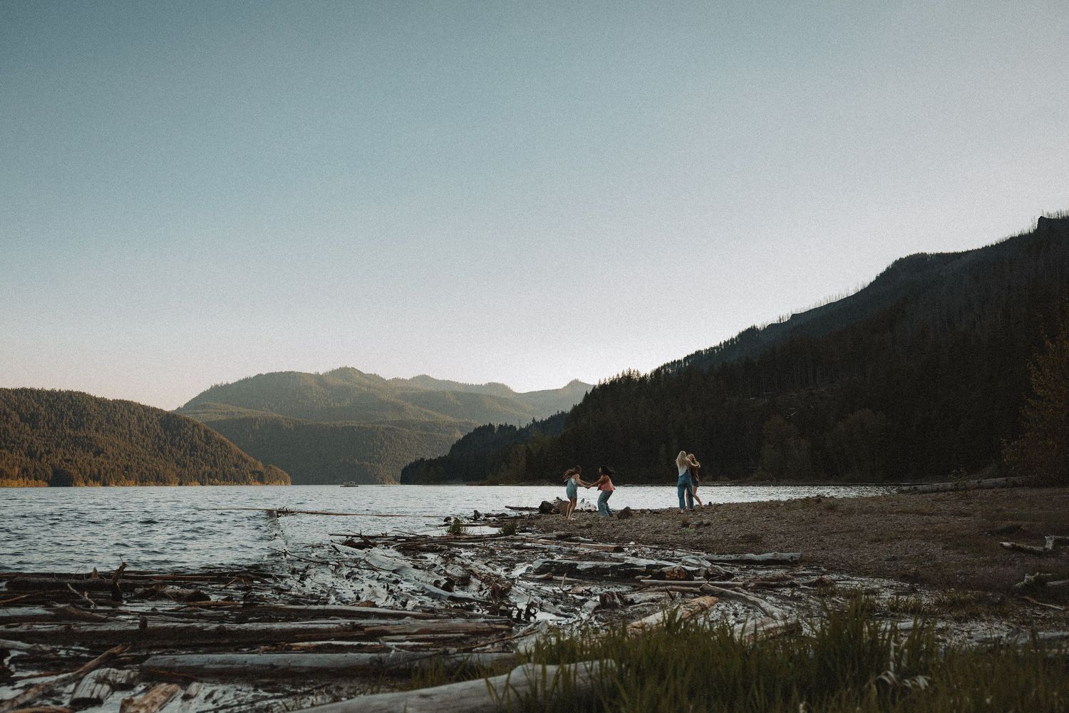 Small group of people gathered on a driftwood-covered mountain lake shoreline at sunset.