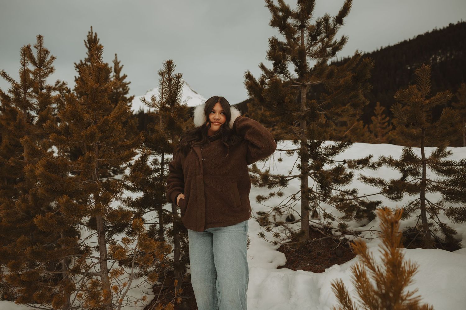 Young woman in brown jacket posing among snow-dusted pine trees in a winter mountain setting.