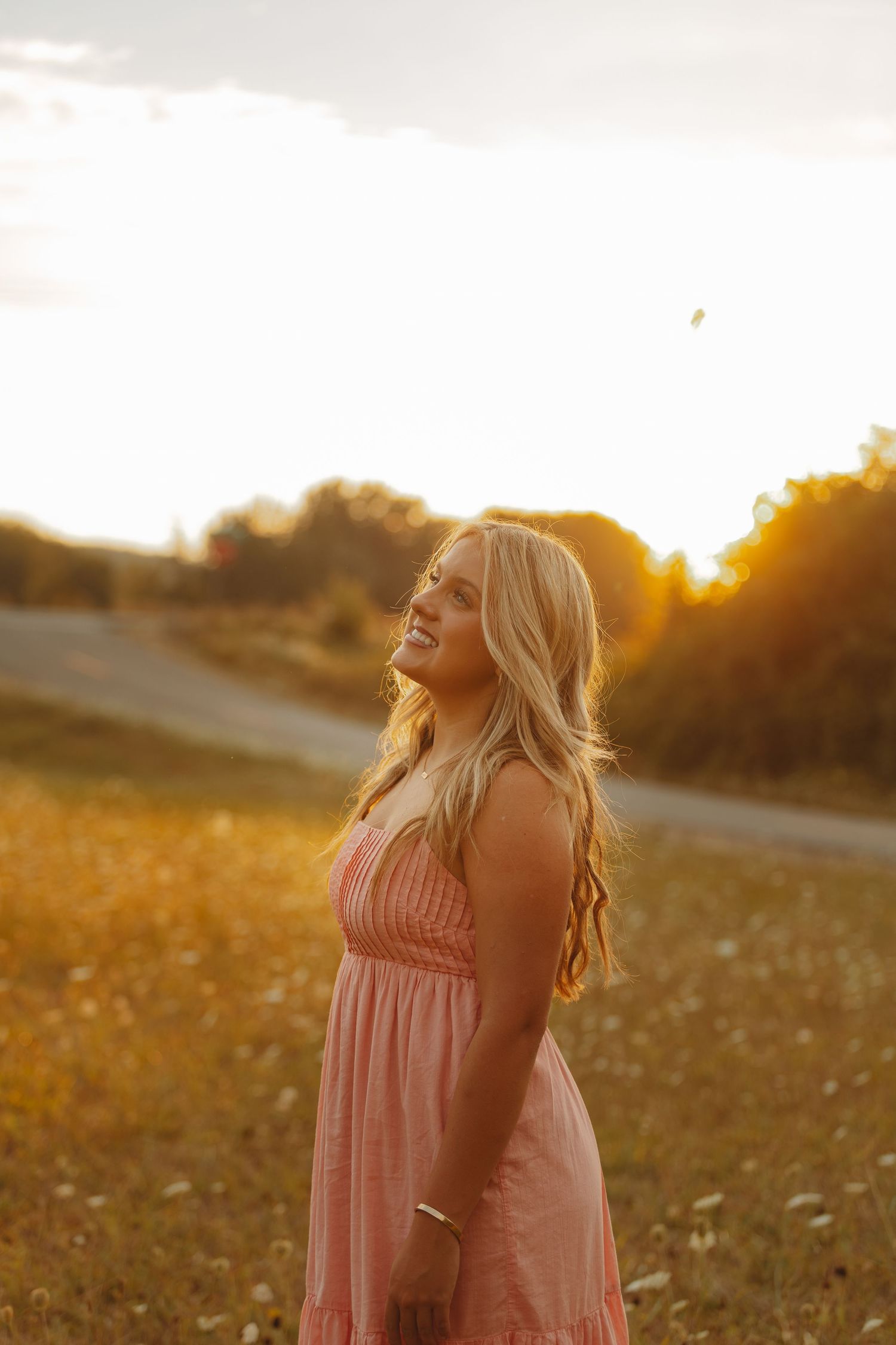 Blonde woman in pink maxi dress laughing in golden field during stunning sunset.