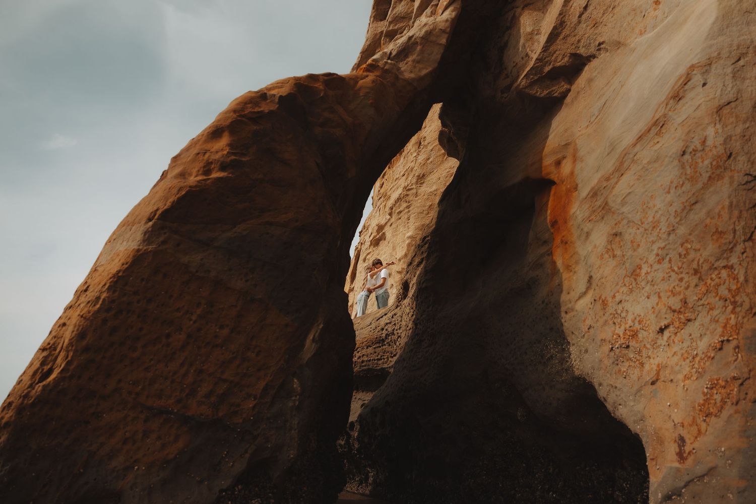 A narrow rock arch formation in red sandstone desert landscape under cloudy sky with a person visible through the opening.