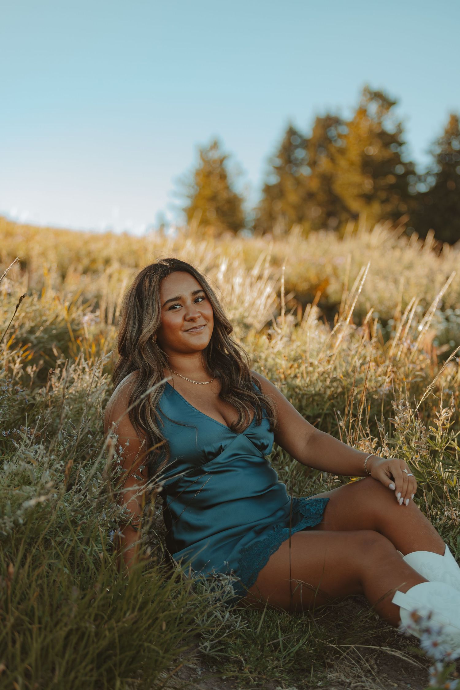 Young woman in teal dress sitting in golden meadow grass at sunset with trees in background.