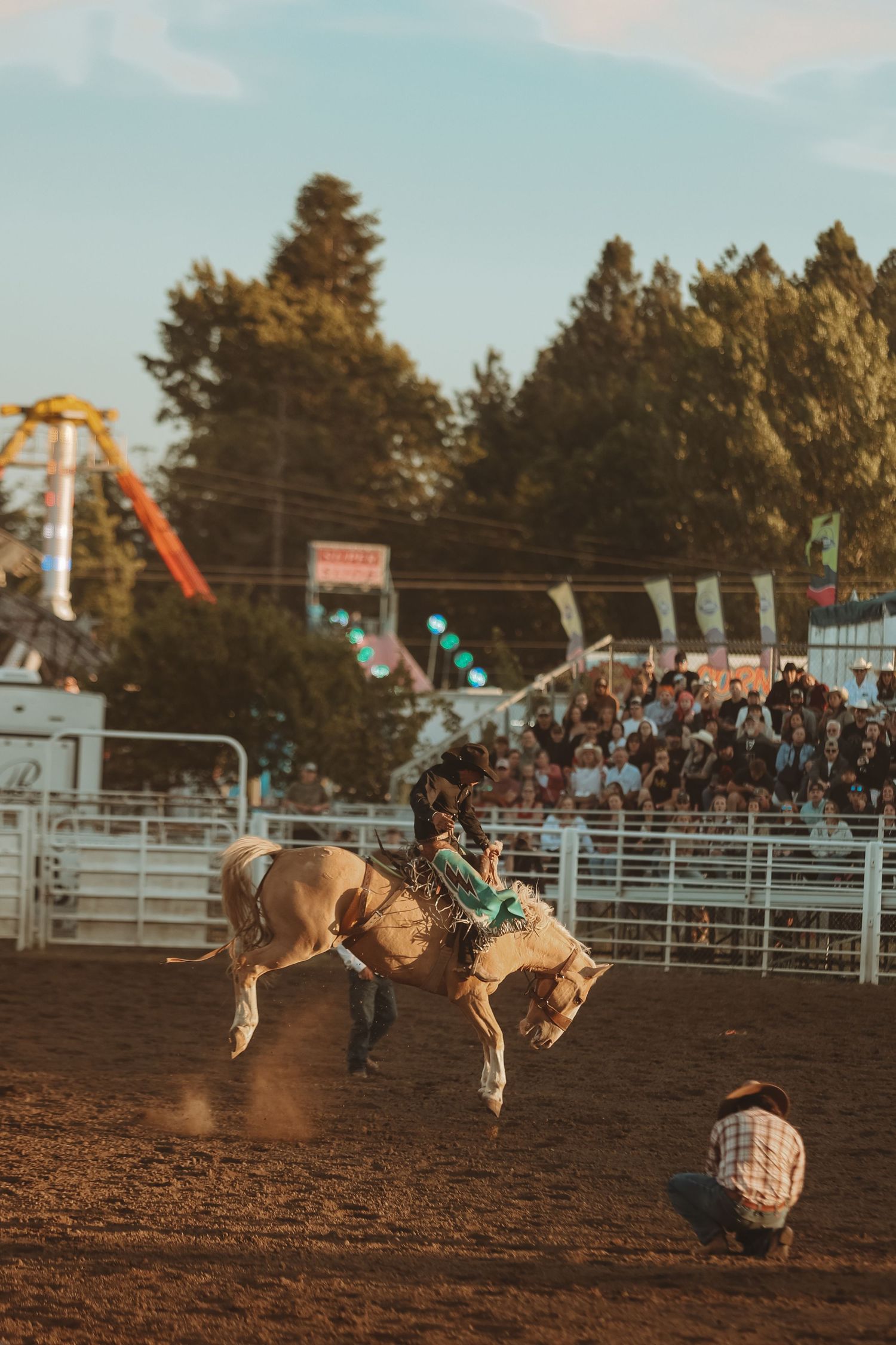 Bronc rider on bucking horse at sunset with carnival rides visible in background.
