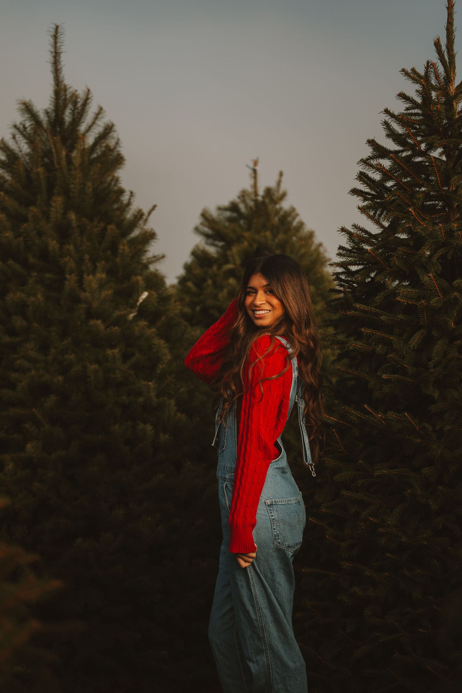 A person in denim overalls and red sweater smiles while standing among Christmas trees at sunset.