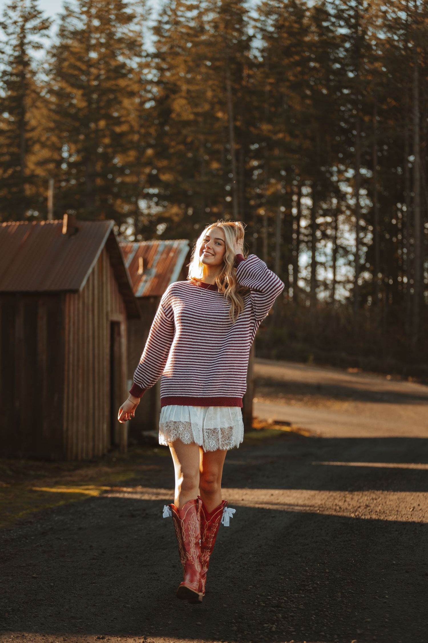Person in striped shirt and boots walks along dirt path near rustic buildings at golden hour.