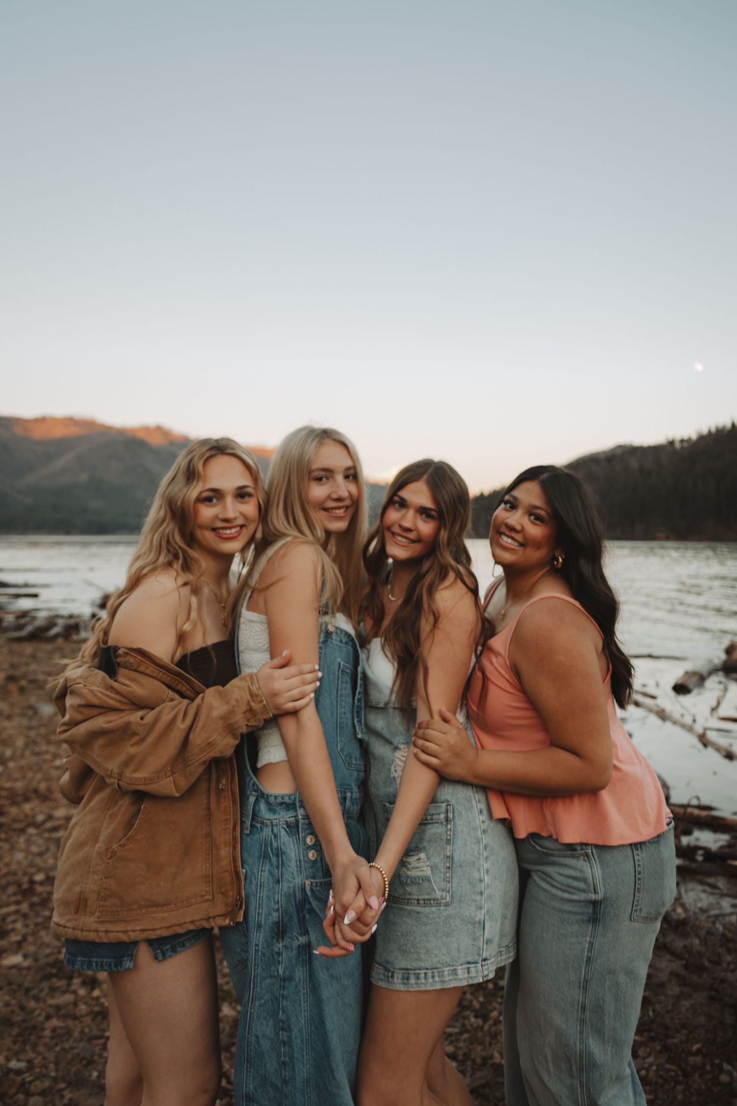Four young women holding hands and smiling by a mountain lake at sunset.