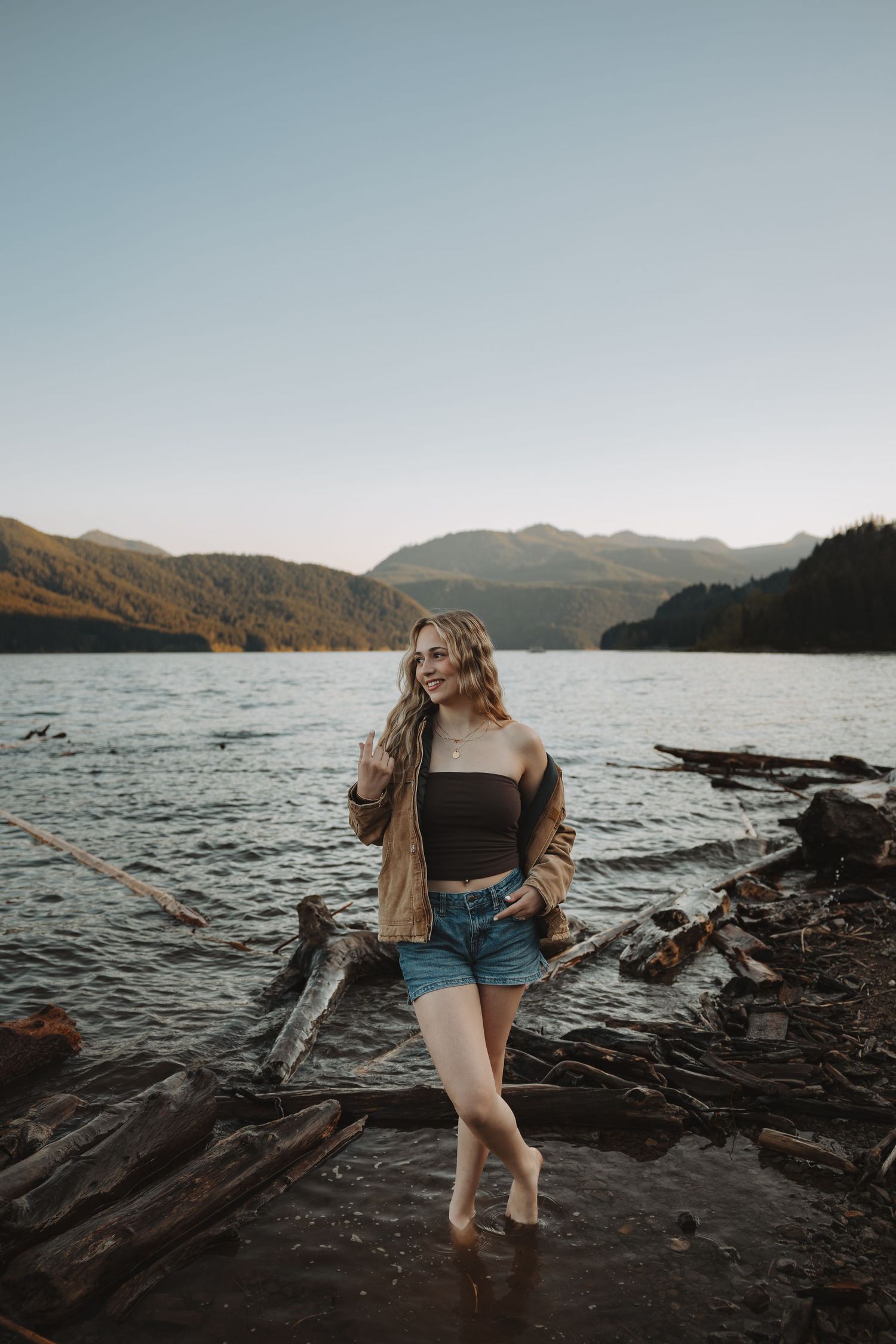 Smiling blonde woman in black crop top and denim shorts stands on driftwood by mountain lake.