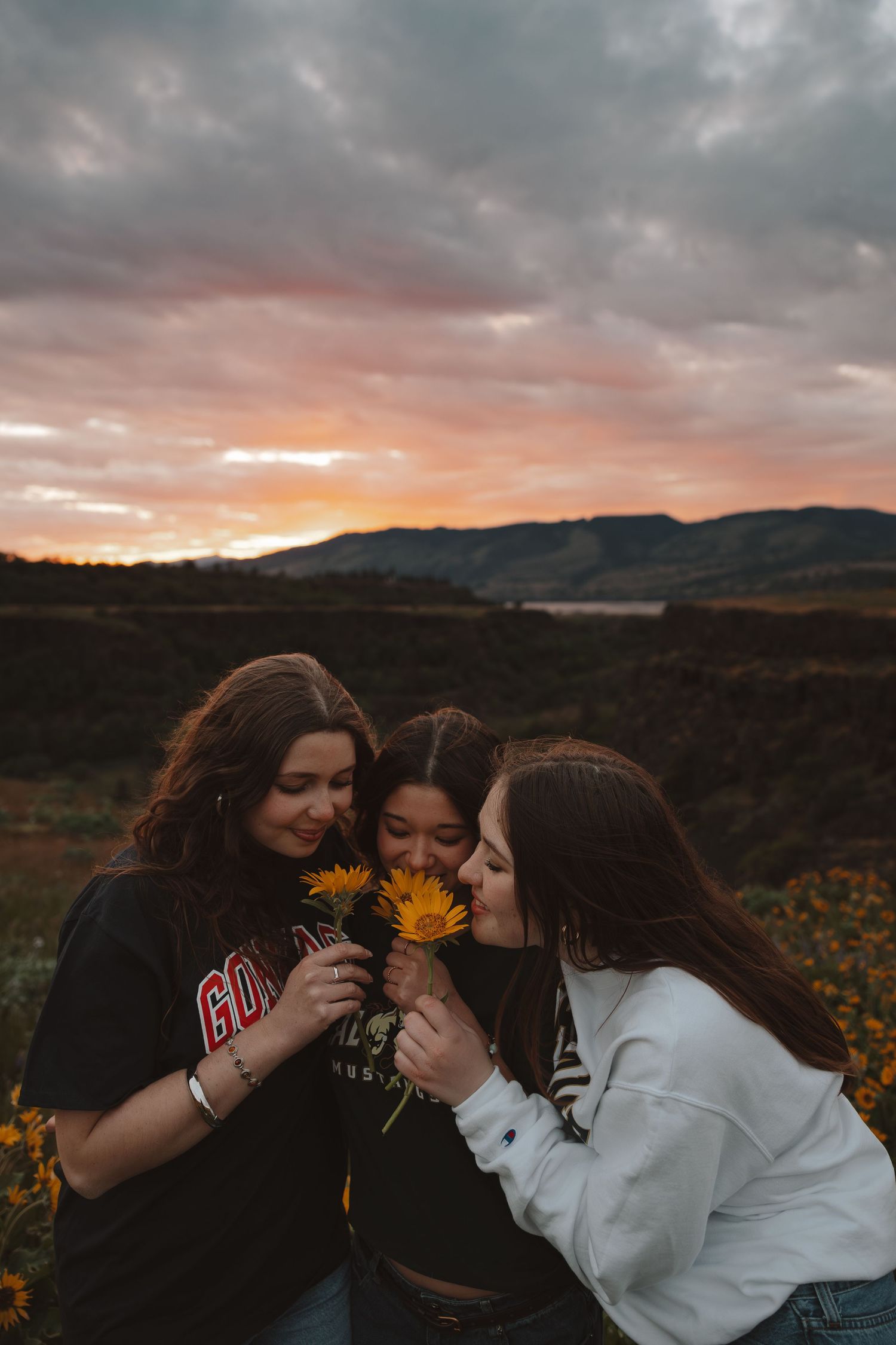Three young women smell a bouquet of yellow wildflowers together during a colorful sunset over mountain landscape.